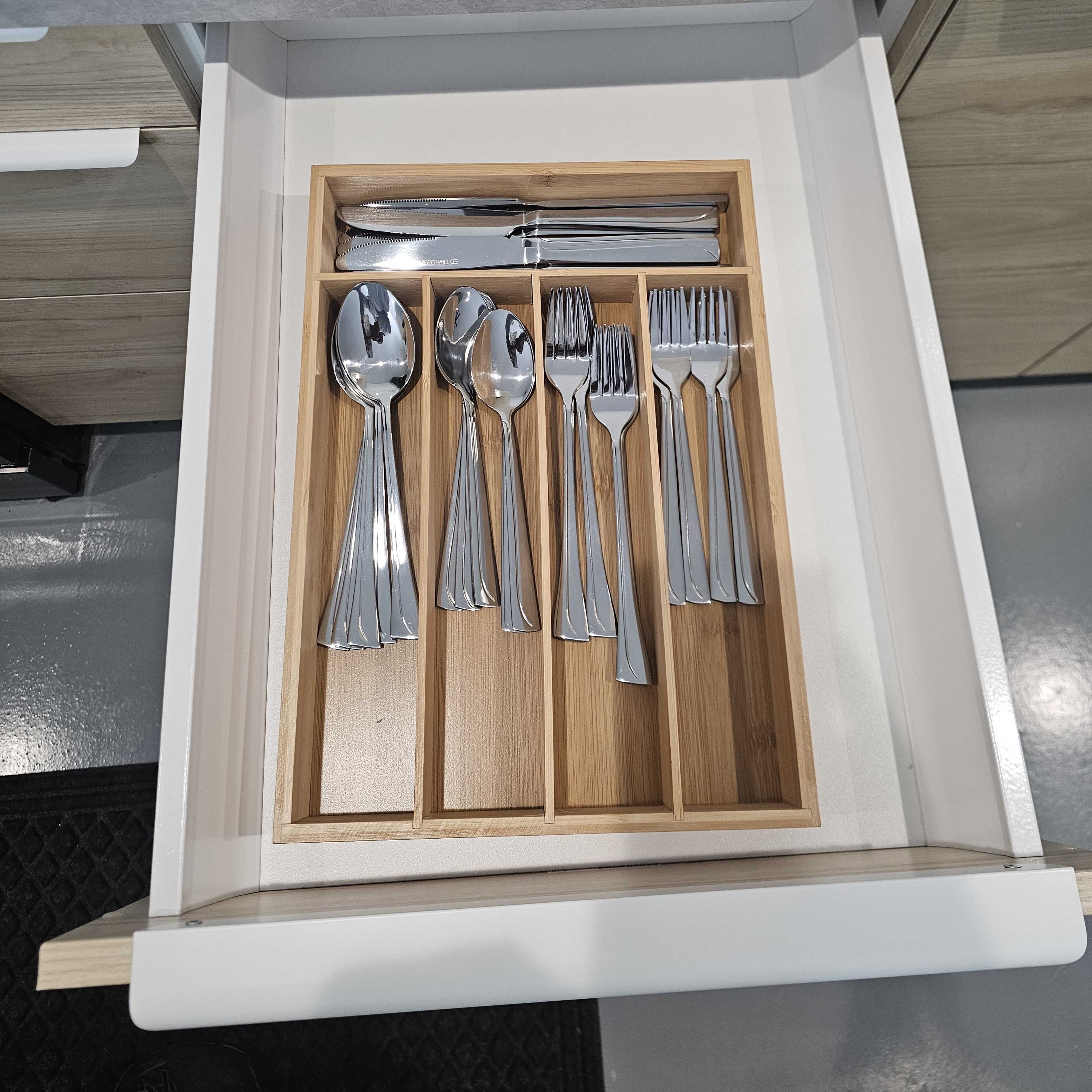 An open kitchen drawer containing a wooden organizer holding various stainless steel cutlery including spoons, forks, and knives.