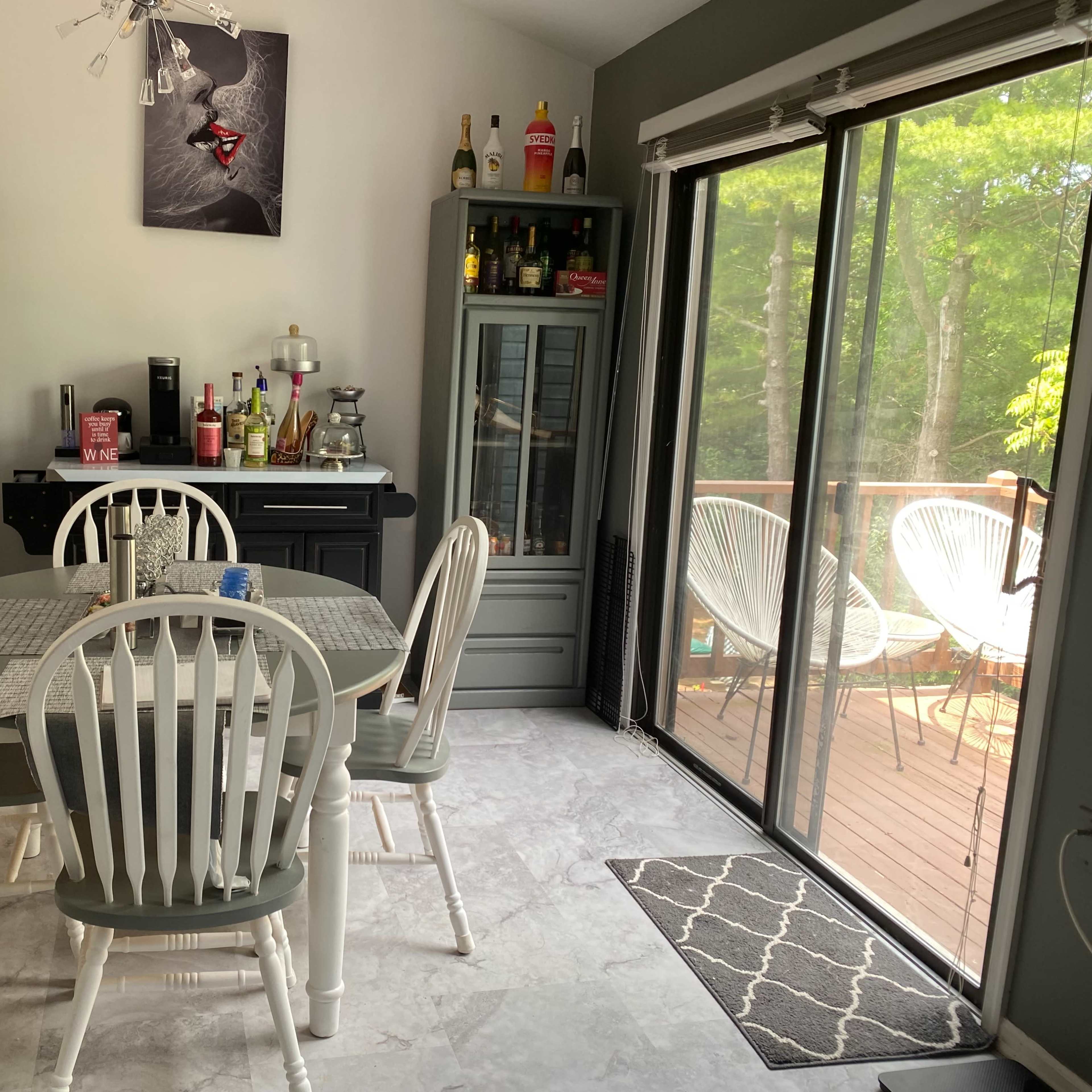 A dining area with a white table and four chairs, flanked by two cabinets filled with bottles, and a large sliding glass door leading to a deck outside.
