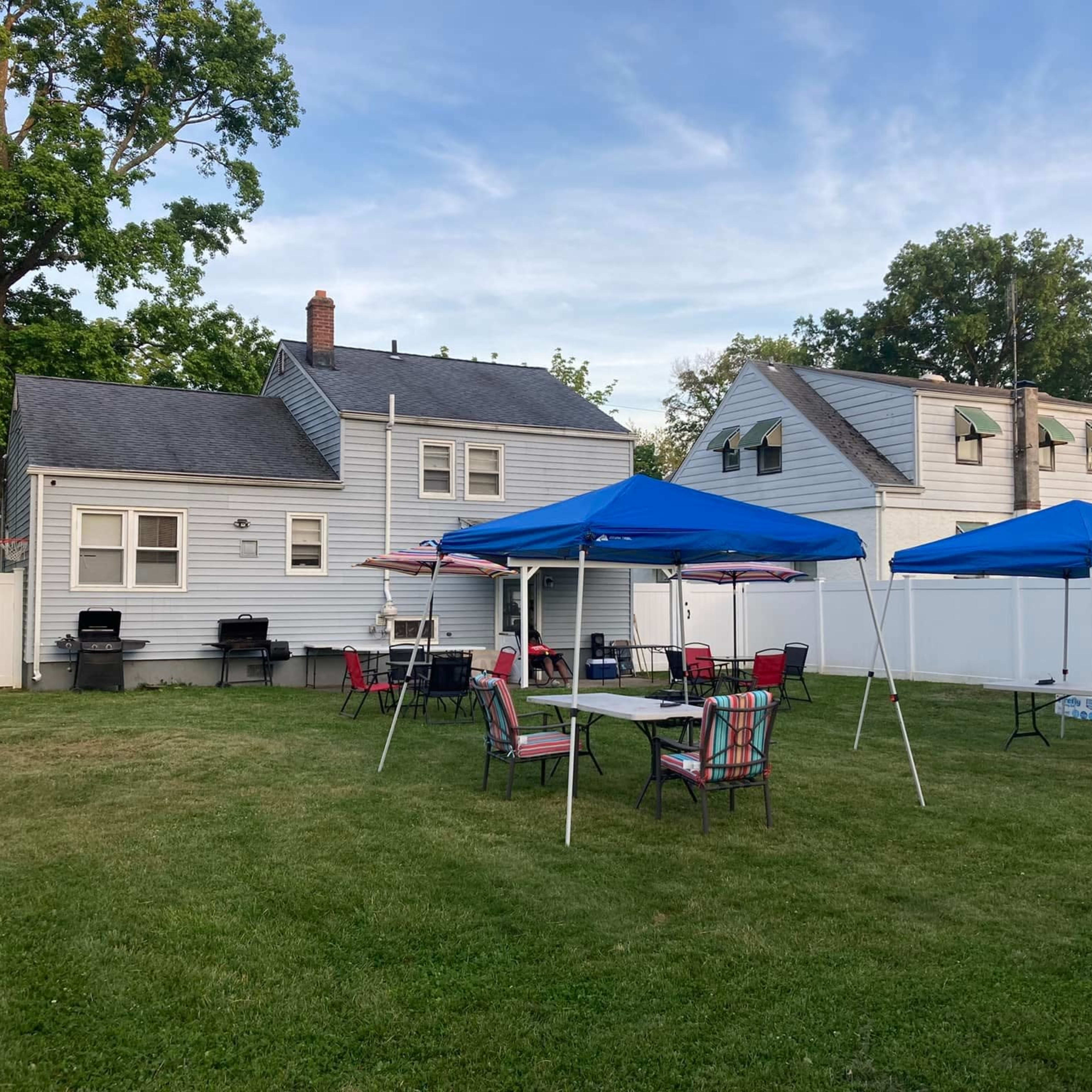A grassy backyard features several blue tents, tables, and chairs set up near a white fence and two houses.