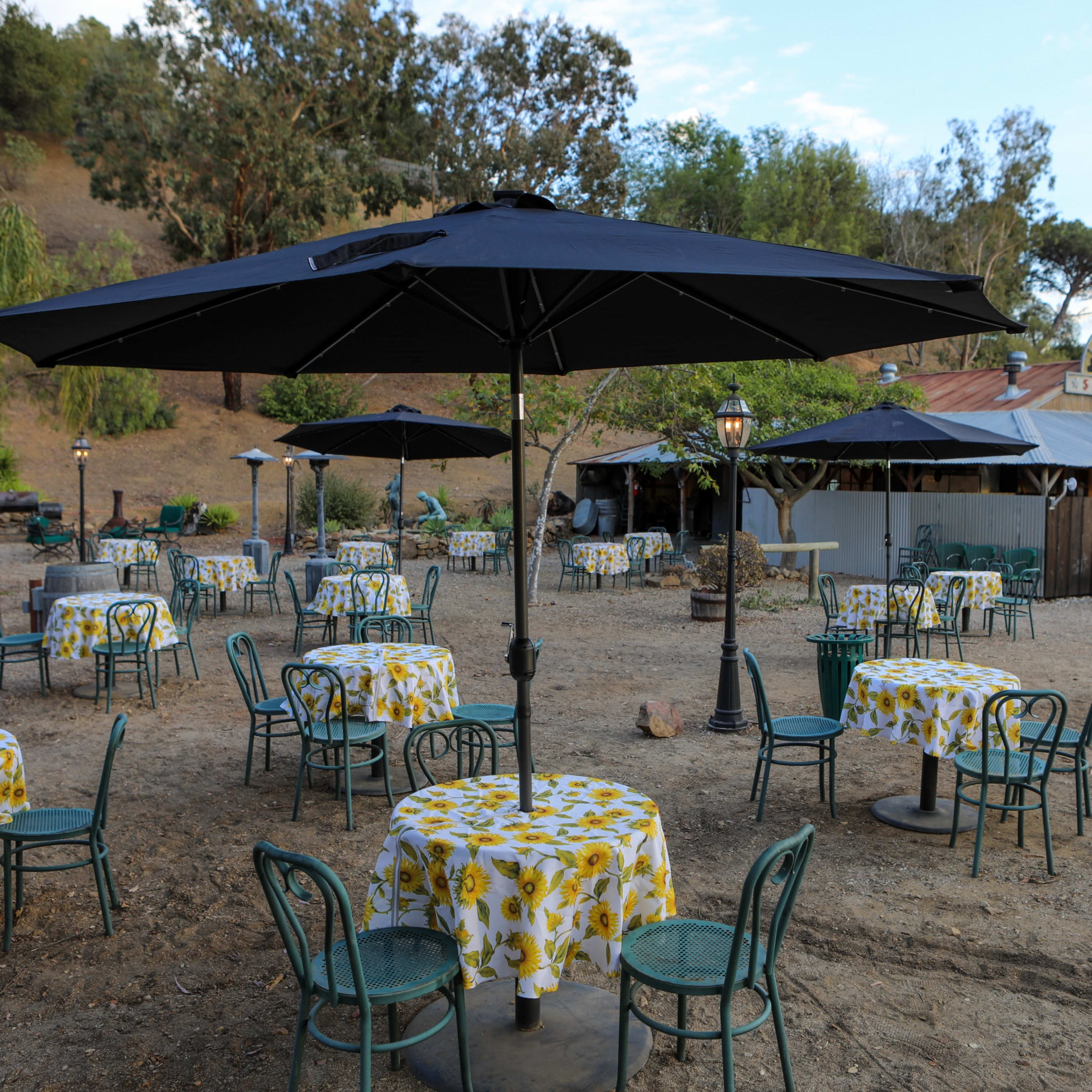 The scene shows an outdoor dining area with several tables covered in yellow floral tablecloths, black umbrellas, and a rustic building in the background surrounded by trees.
