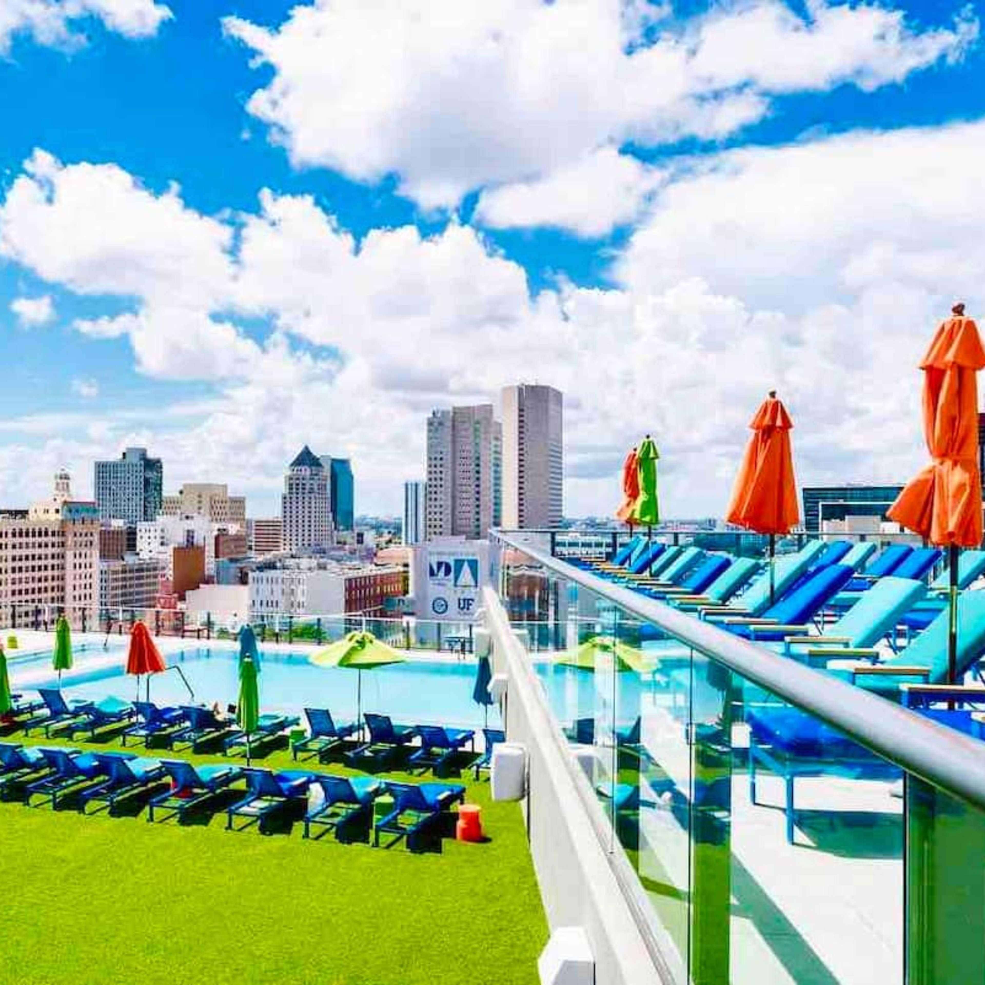 A rooftop pool area with lounge chairs and umbrellas overlooking a city skyline under a partly cloudy sky.