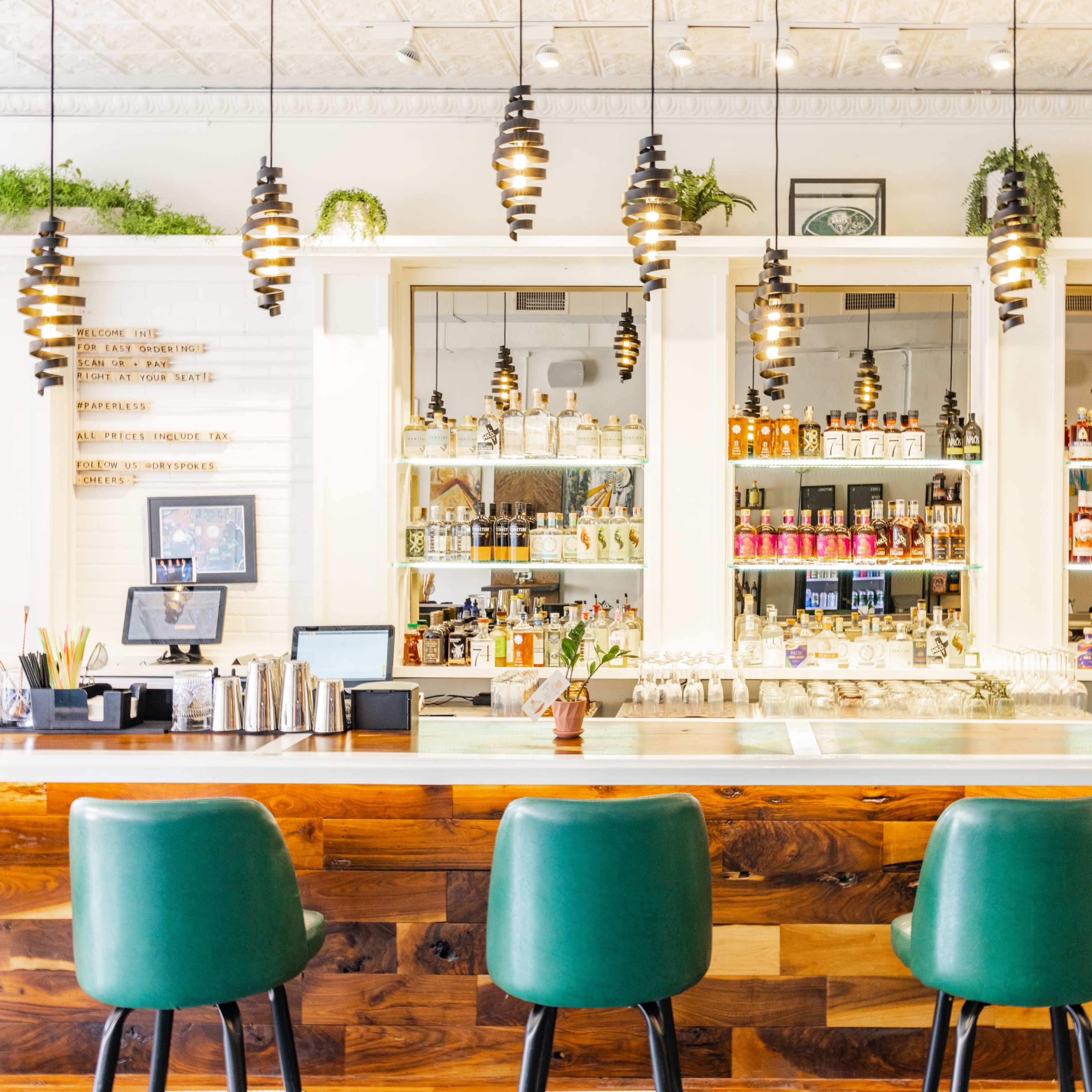 A well-lit bar with a wooden counter, green upholstered stools, and shelves displaying various bottles of alcohol.