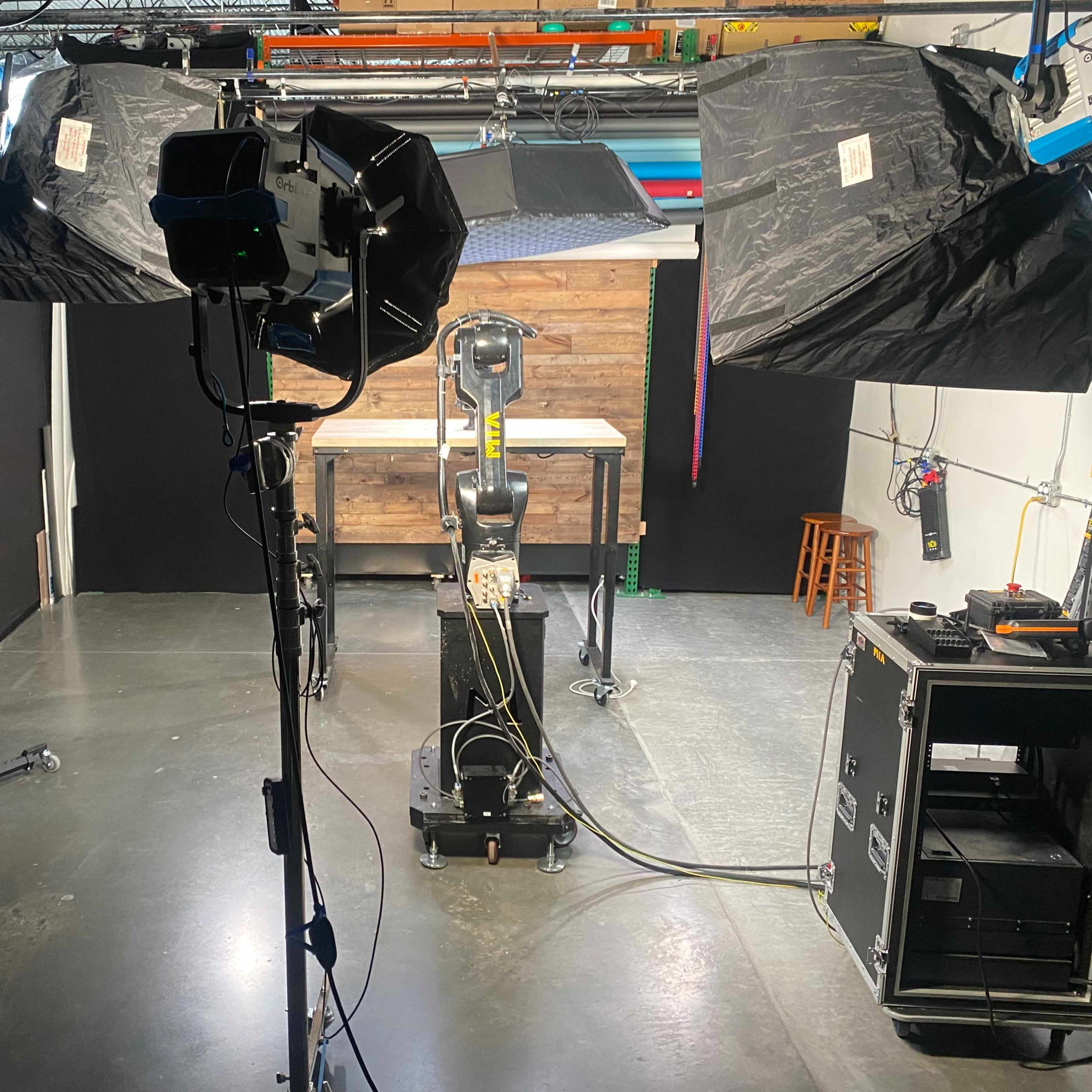 A studio setup with two large softbox lights, a robotic arm in the center, and various equipment arranged around a wooden tabletop in a black-walled space.