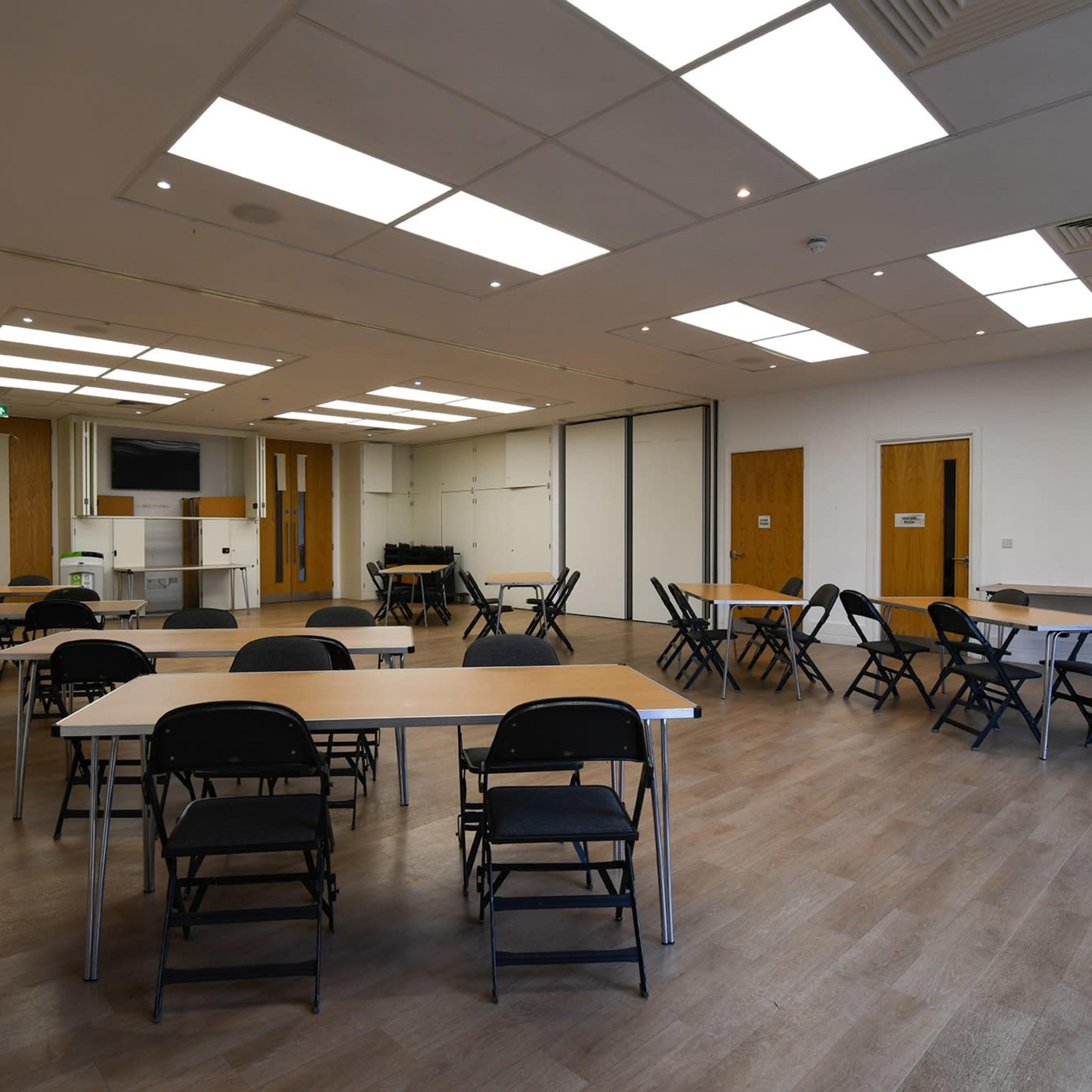 A spacious meeting room features several tables and black chairs arranged throughout the area, with large windows and bright overhead lights.