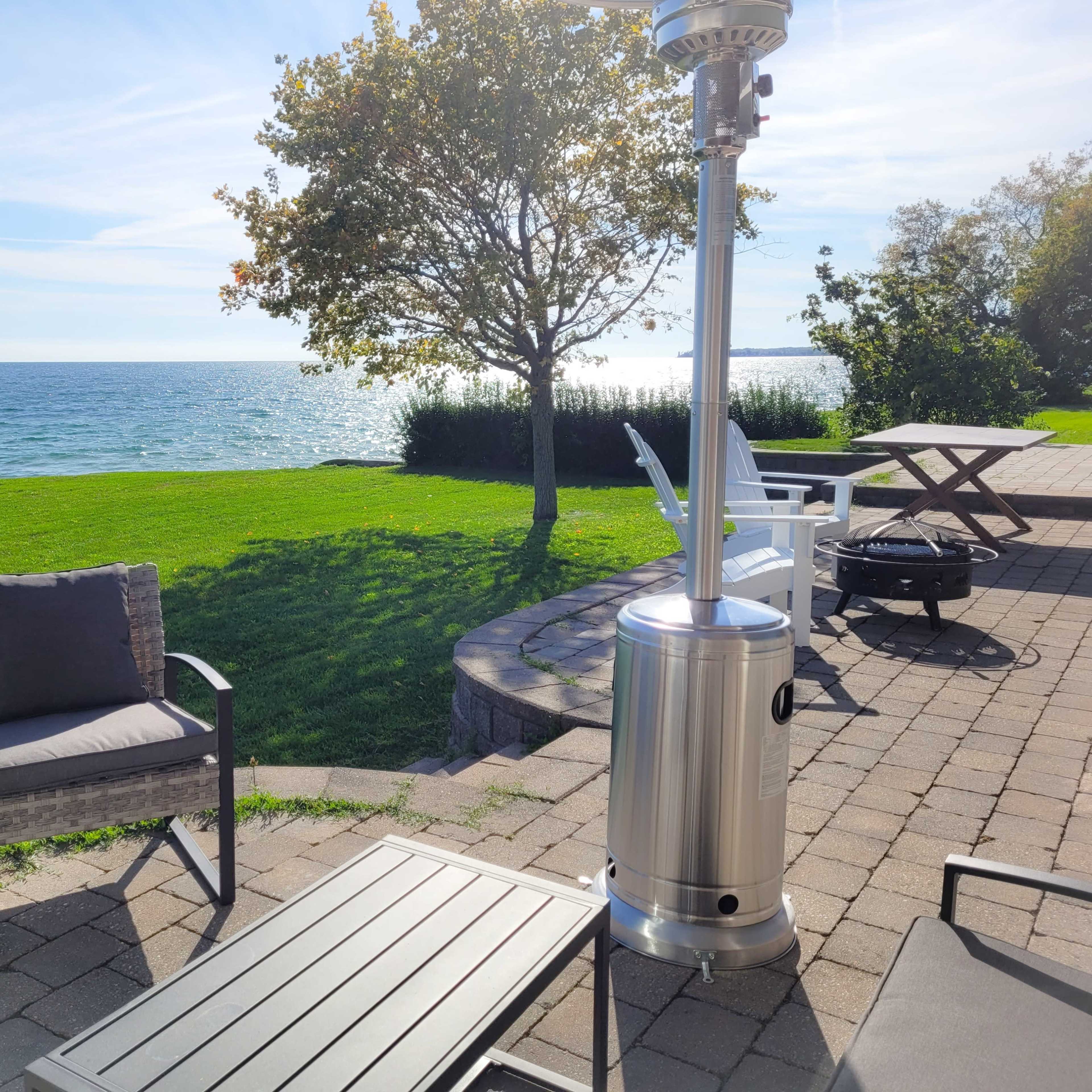 A patio setup features a metal heater and seating arranged near a lake with grassy areas and a tree in the background.