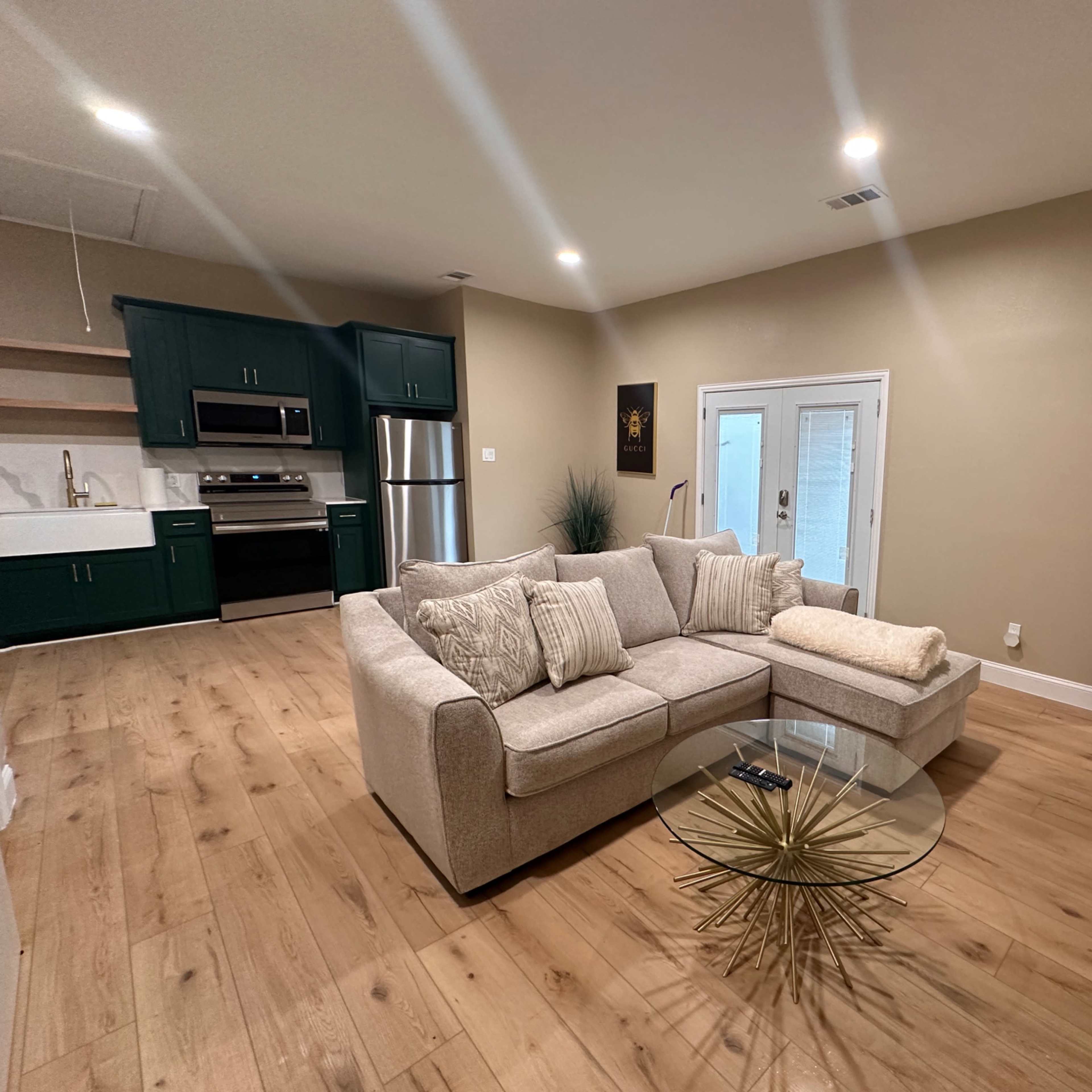 The living space features a light-colored sectional sofa, a glass coffee table with a gold starburst base, and a kitchen area with dark green cabinetry.