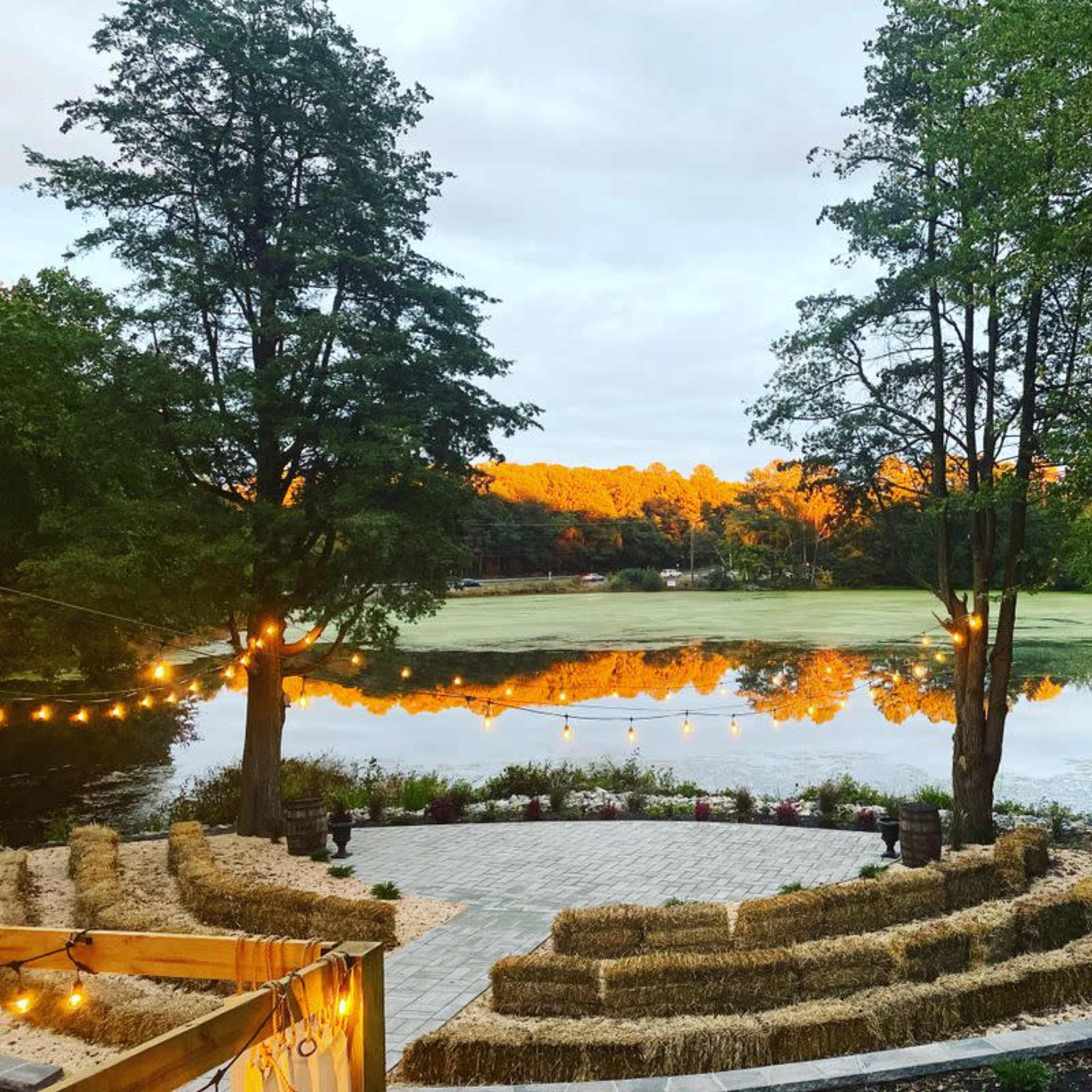 A circular seating area made of hay bales overlooks a tranquil lake bordered by trees, reflecting the orange tones of the sunset.