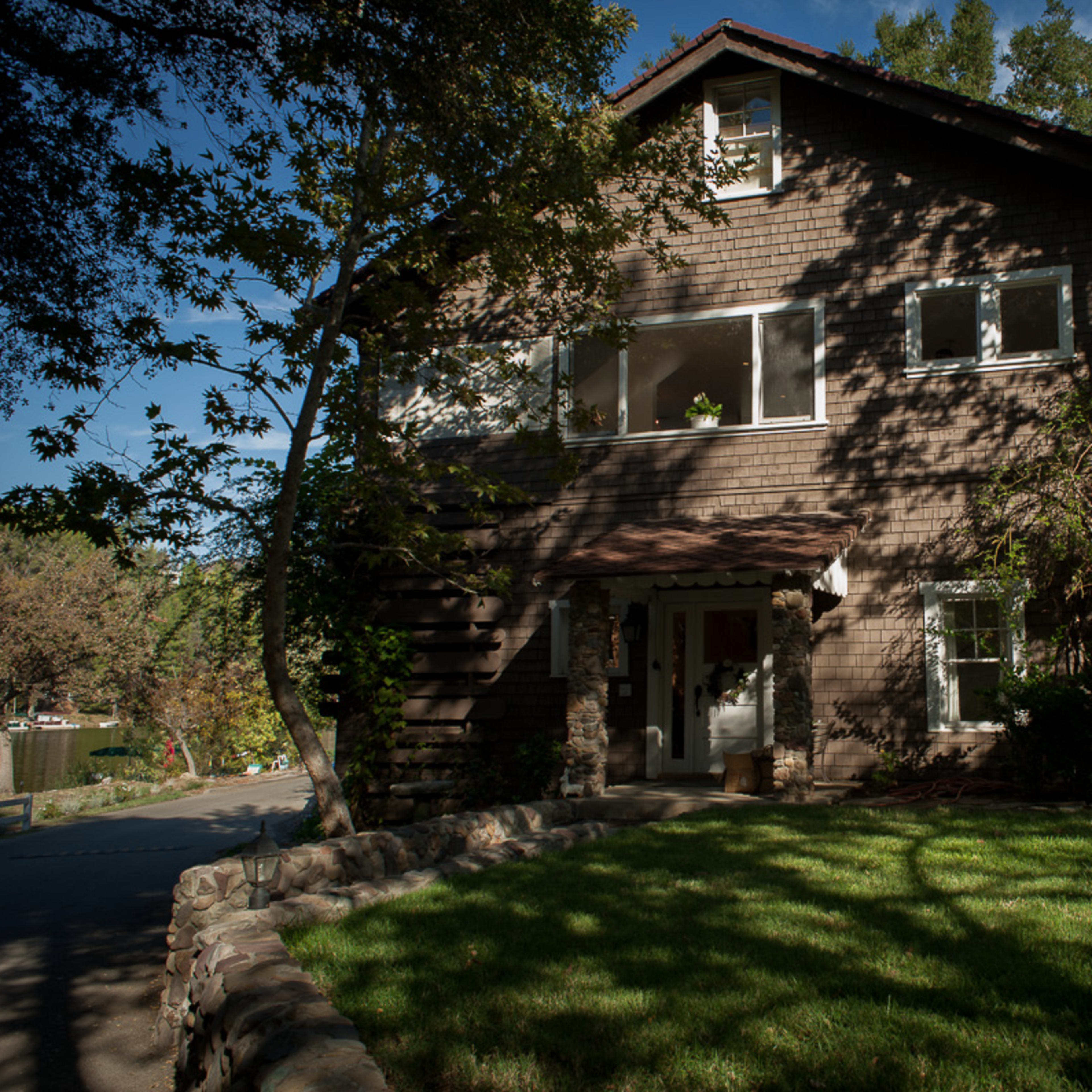 A two-story wooden house with large windows and a garden sits under the shade of trees.