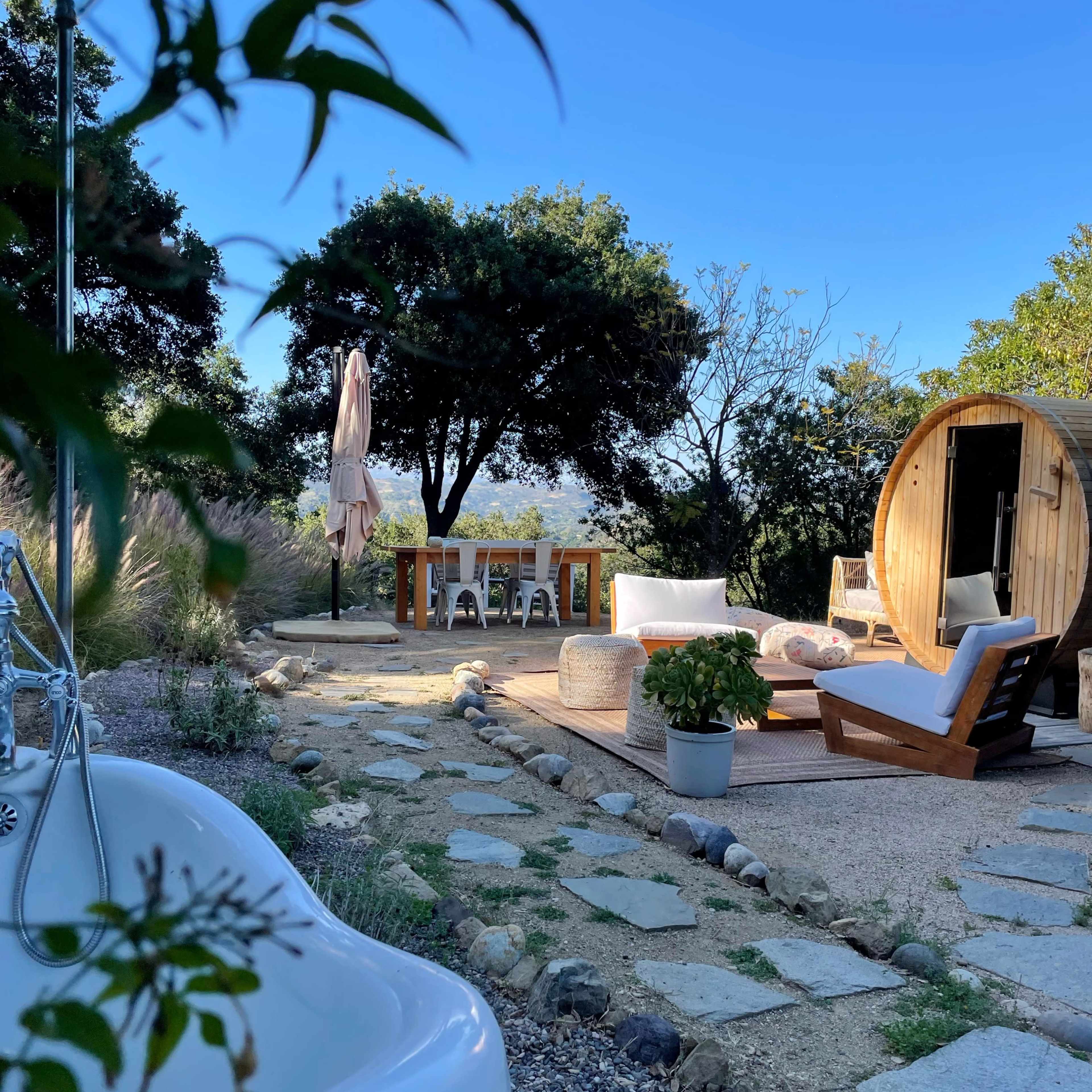 A wooden seating area and a dining table are set up outdoors near a bathtub and a barrel-shaped structure, all surrounded by trees and a clear blue sky.
