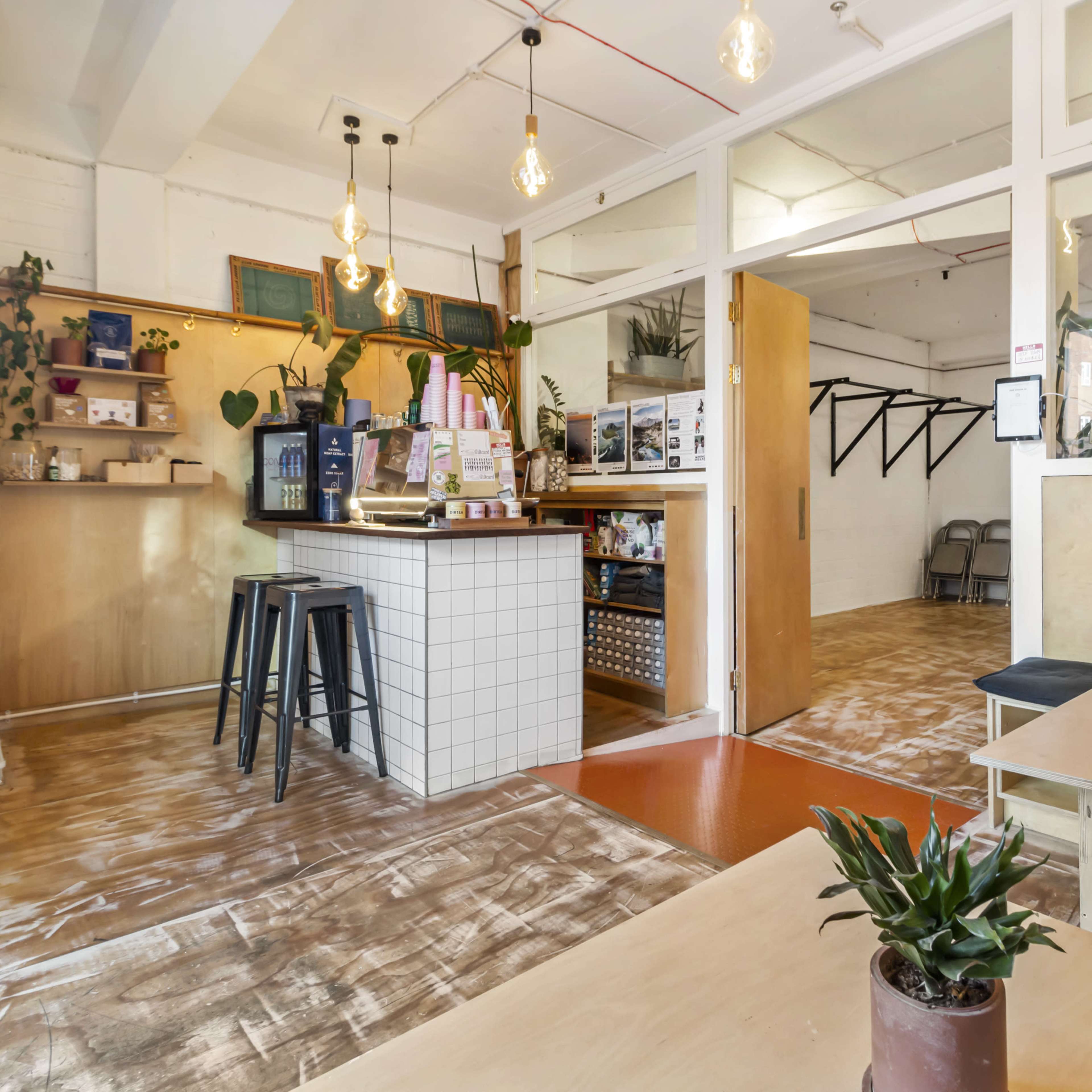 A modern café interior featuring wooden tables, a barista counter with a coffee machine, and green plants in decorative pots.