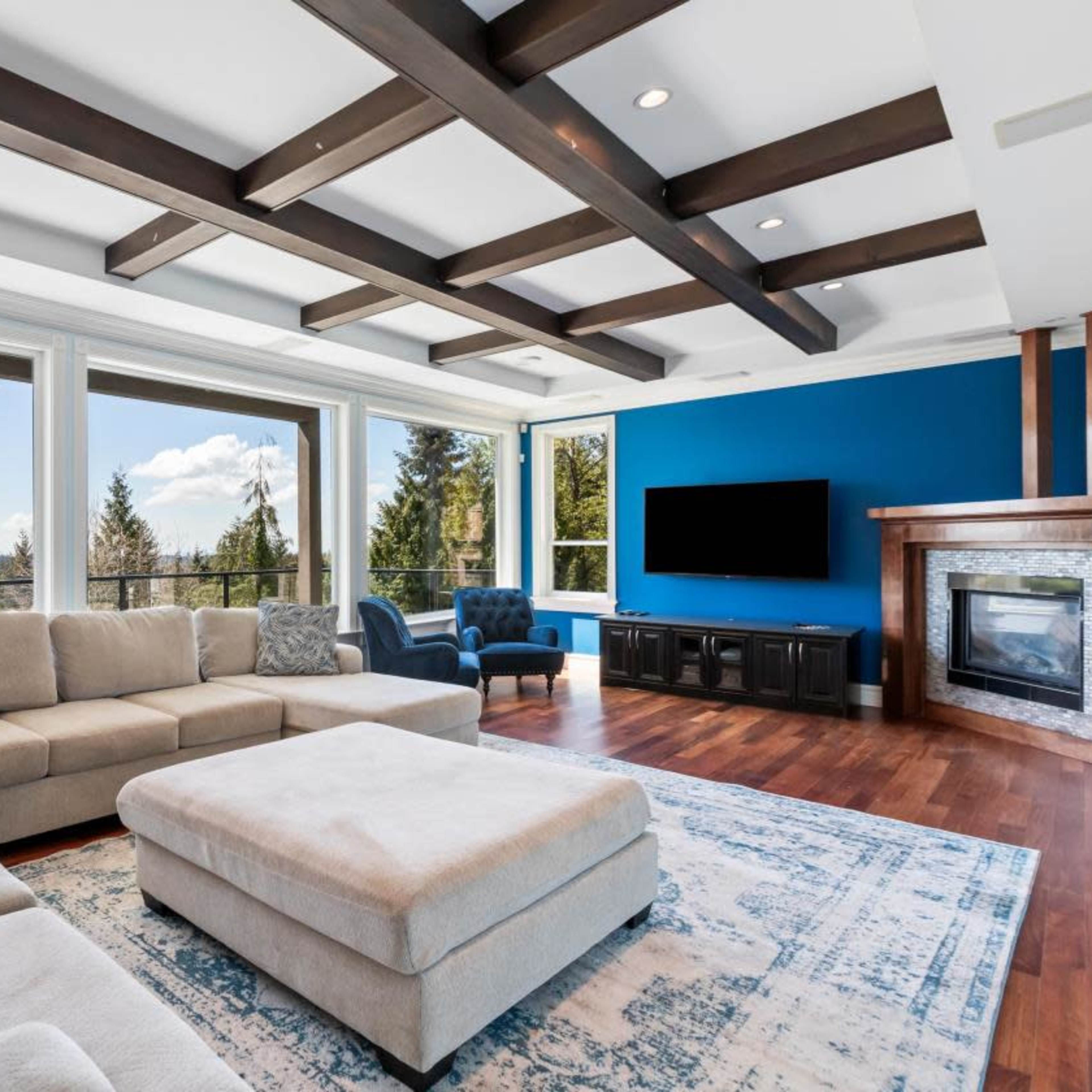 A spacious living room features a large sectional sofa, a patterned rug, and a blue accent wall, with large windows offering views of trees and the sky.