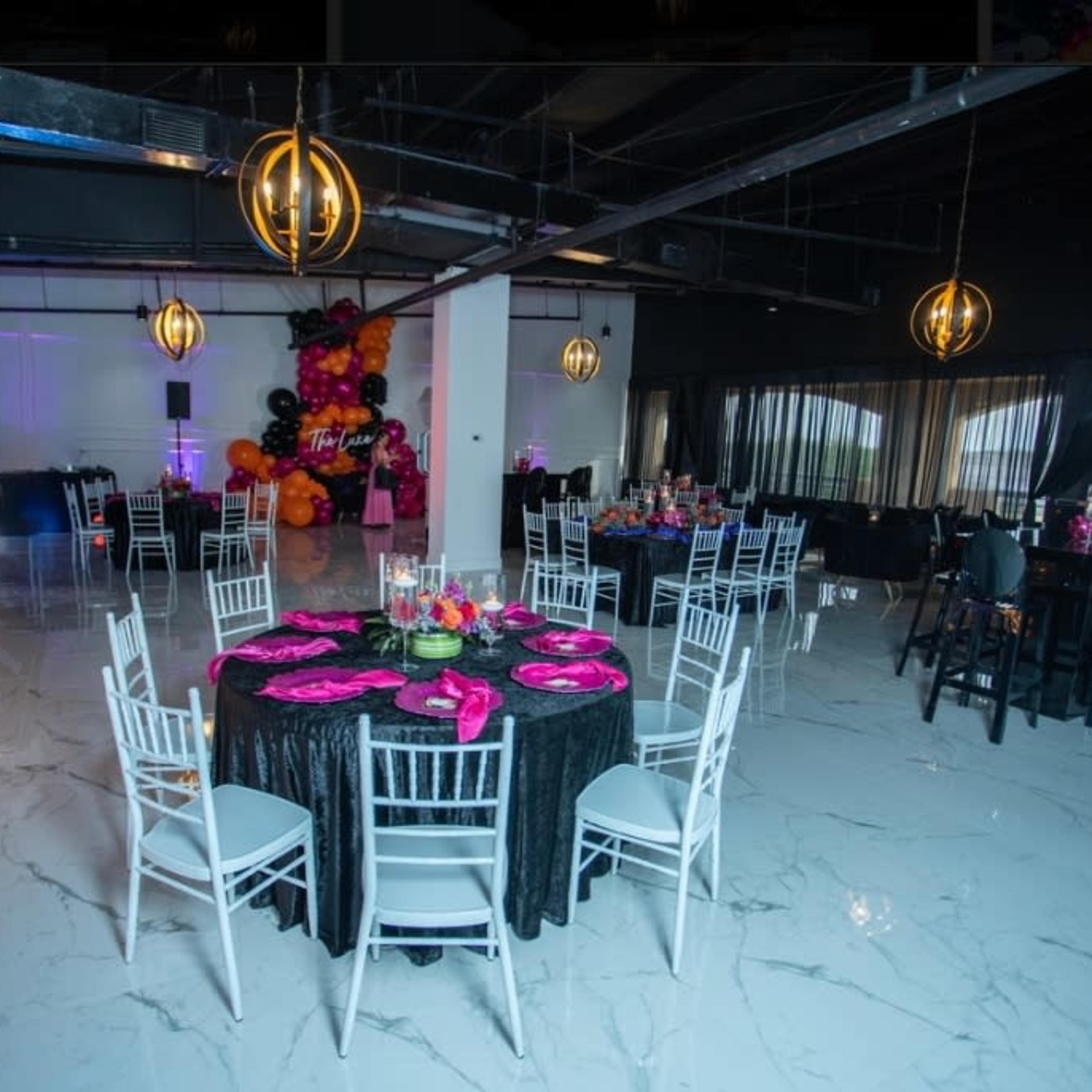 A spacious event hall with black and pink decor, featuring round tables set with pink tableware and colorful floral arrangements, and a backdrop of balloons.