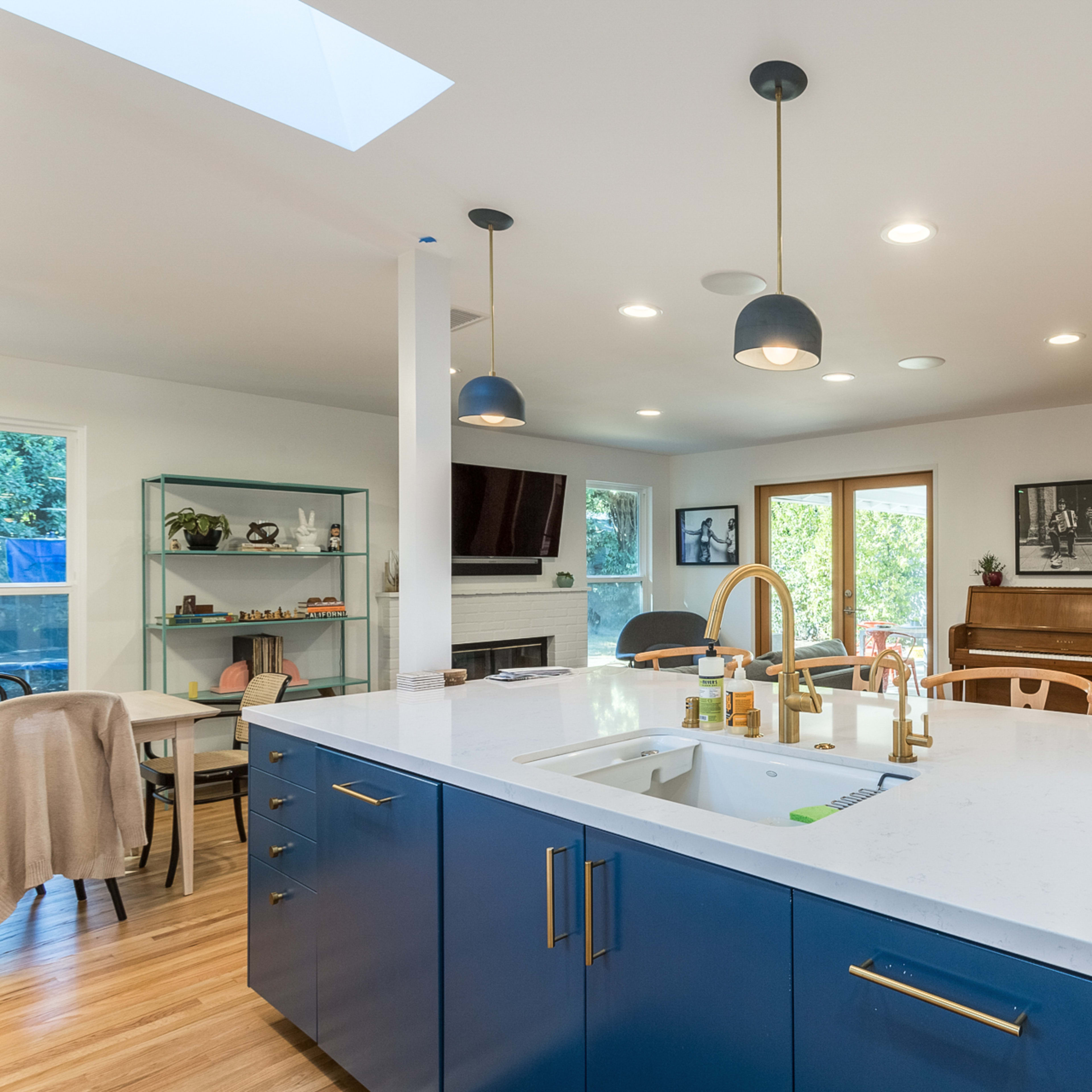 A modern kitchen with a central island, blue cabinetry, a white countertop, and a dining area featuring a piano and large windows.