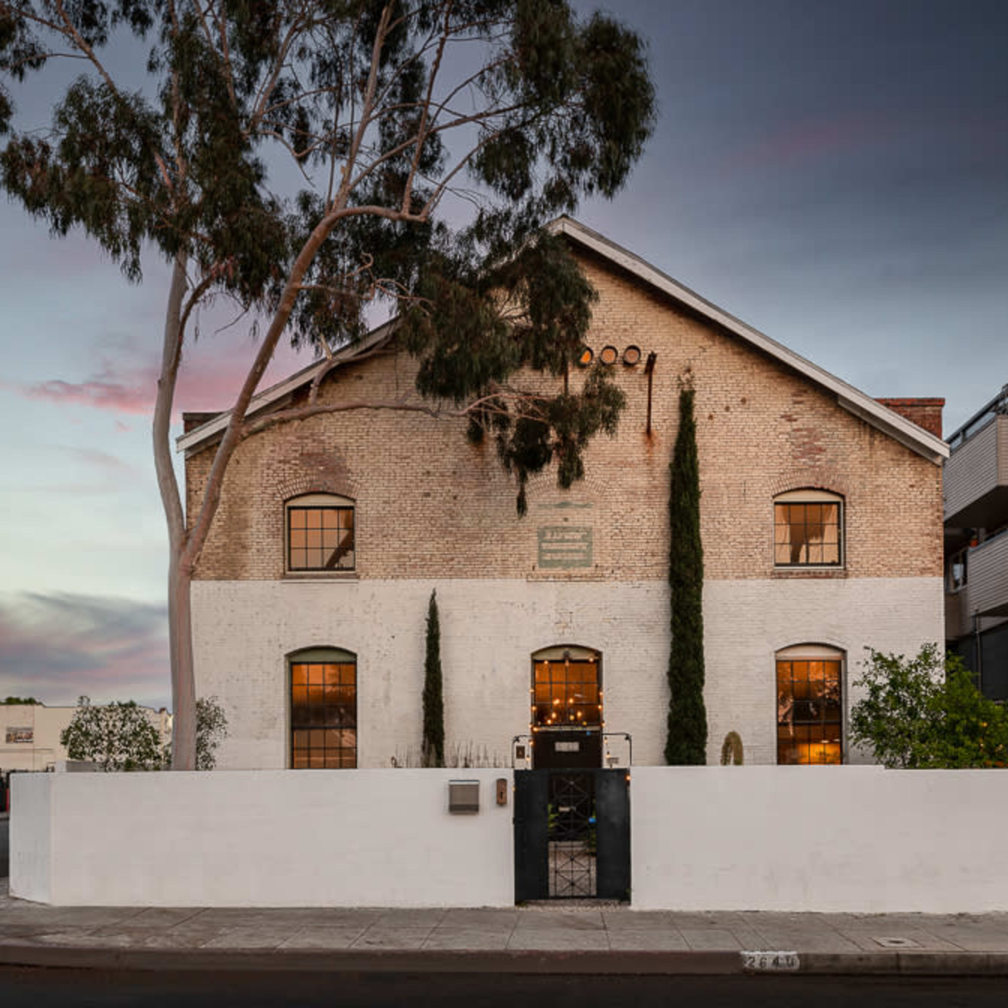 A large, white-walled building with a distinctive, sloped roof, surrounded by trees and other structures on a street corner at dusk.