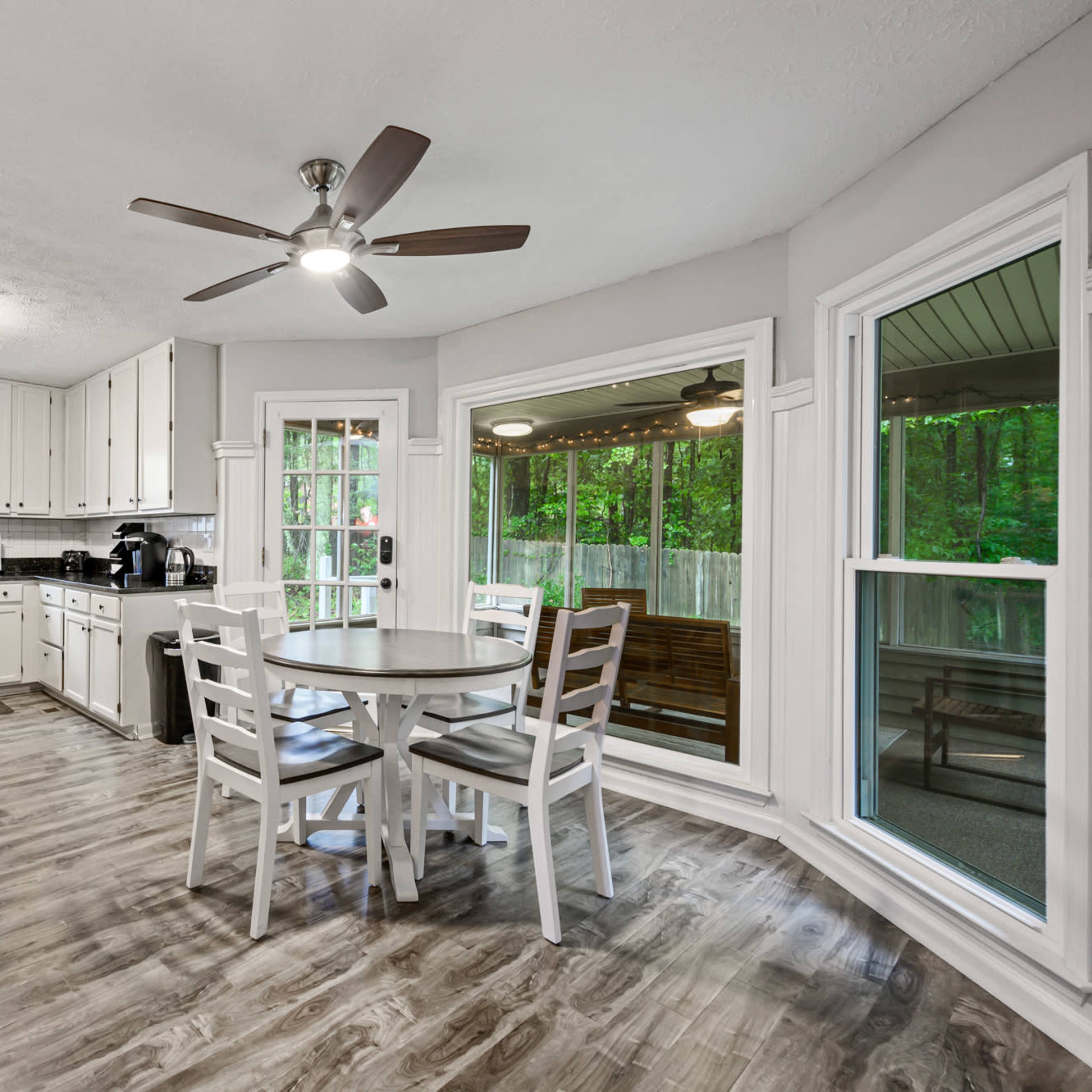 A bright, modern kitchen with white cabinets, a round wooden table, and large windows overlooking a green outdoor area.