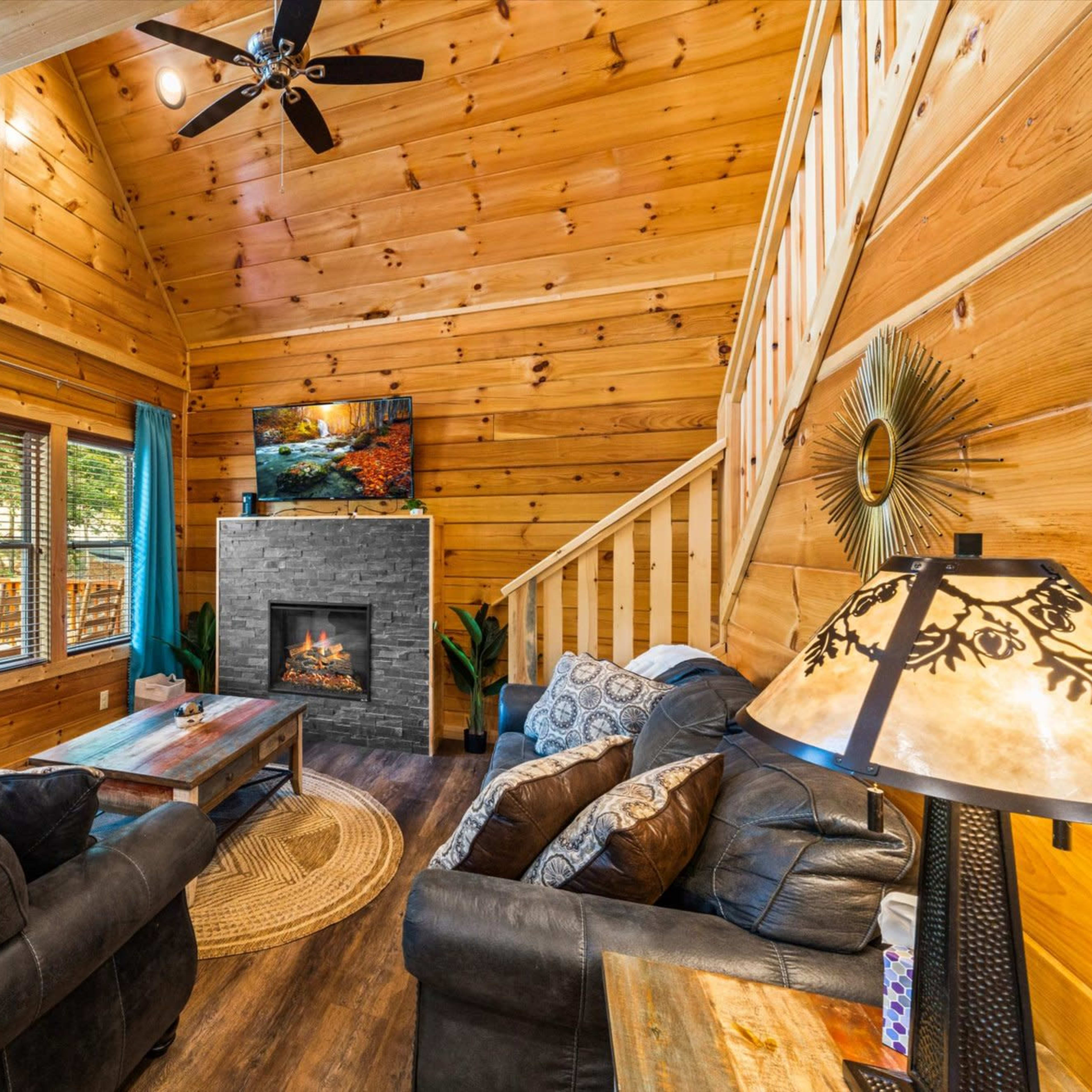 A cozy wooden cabin living room features a stone fireplace, two sofas, and large windows with blue curtains.