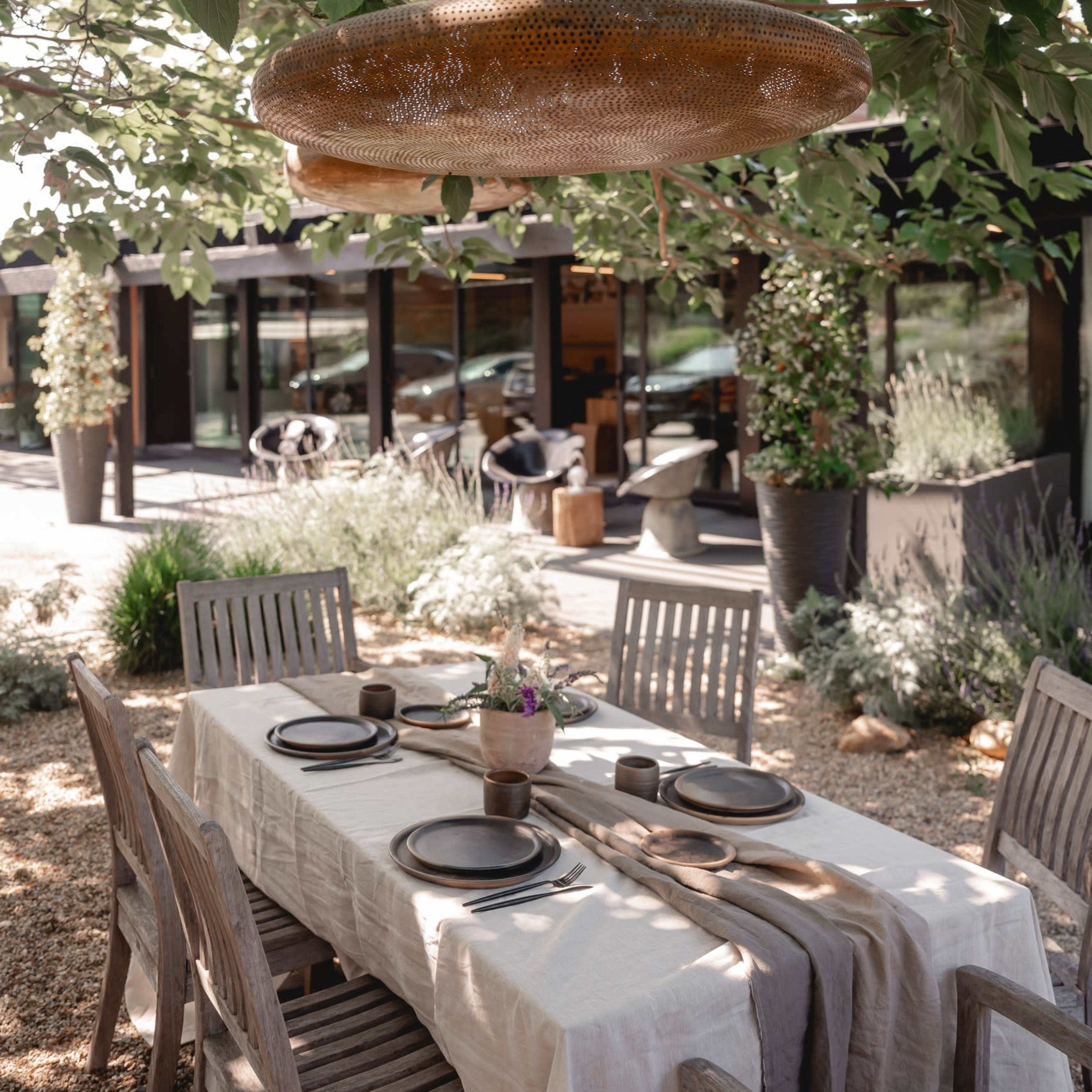 A wooden dining table is set under a tree with hanging light fixtures, surrounded by plants and a modern building in the background.