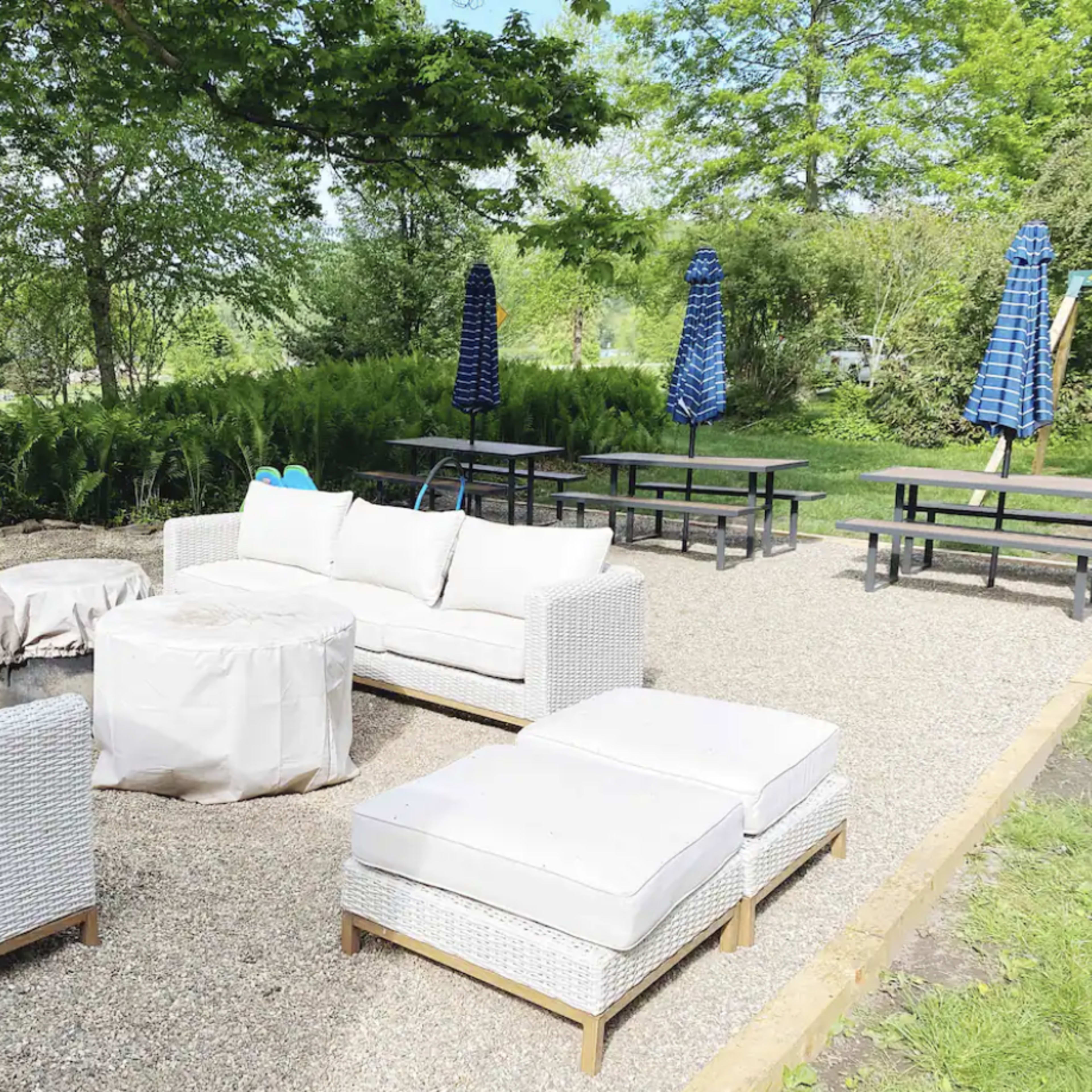 The scene features an outdoor seating area with a white wicker sofa set, a round coffee table, and several picnic tables, surrounded by greenery and blue umbrellas.