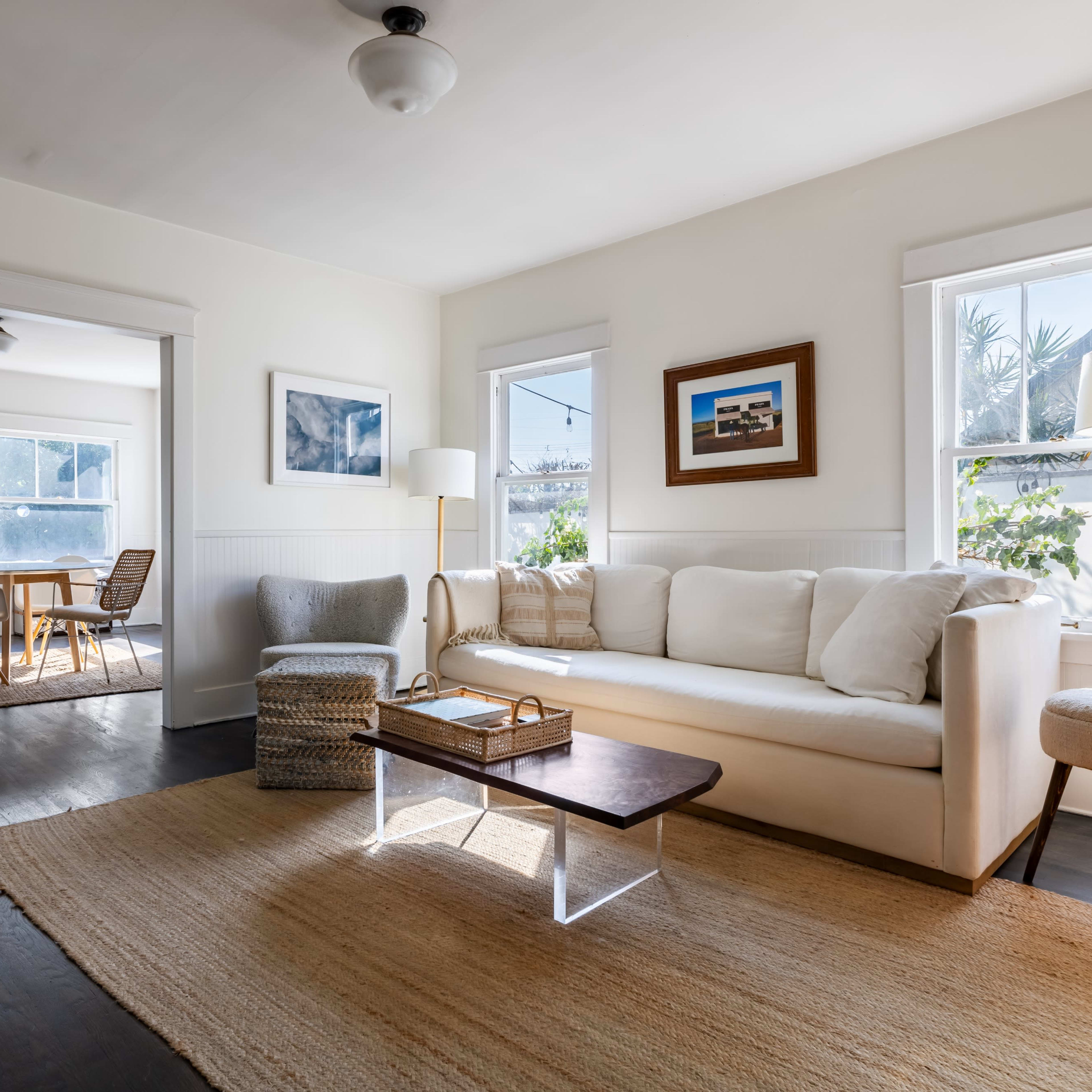 A bright living room features a beige sofa, a wooden coffee table, and large windows that let in natural light, with an adjacent dining area visible through an open doorway.