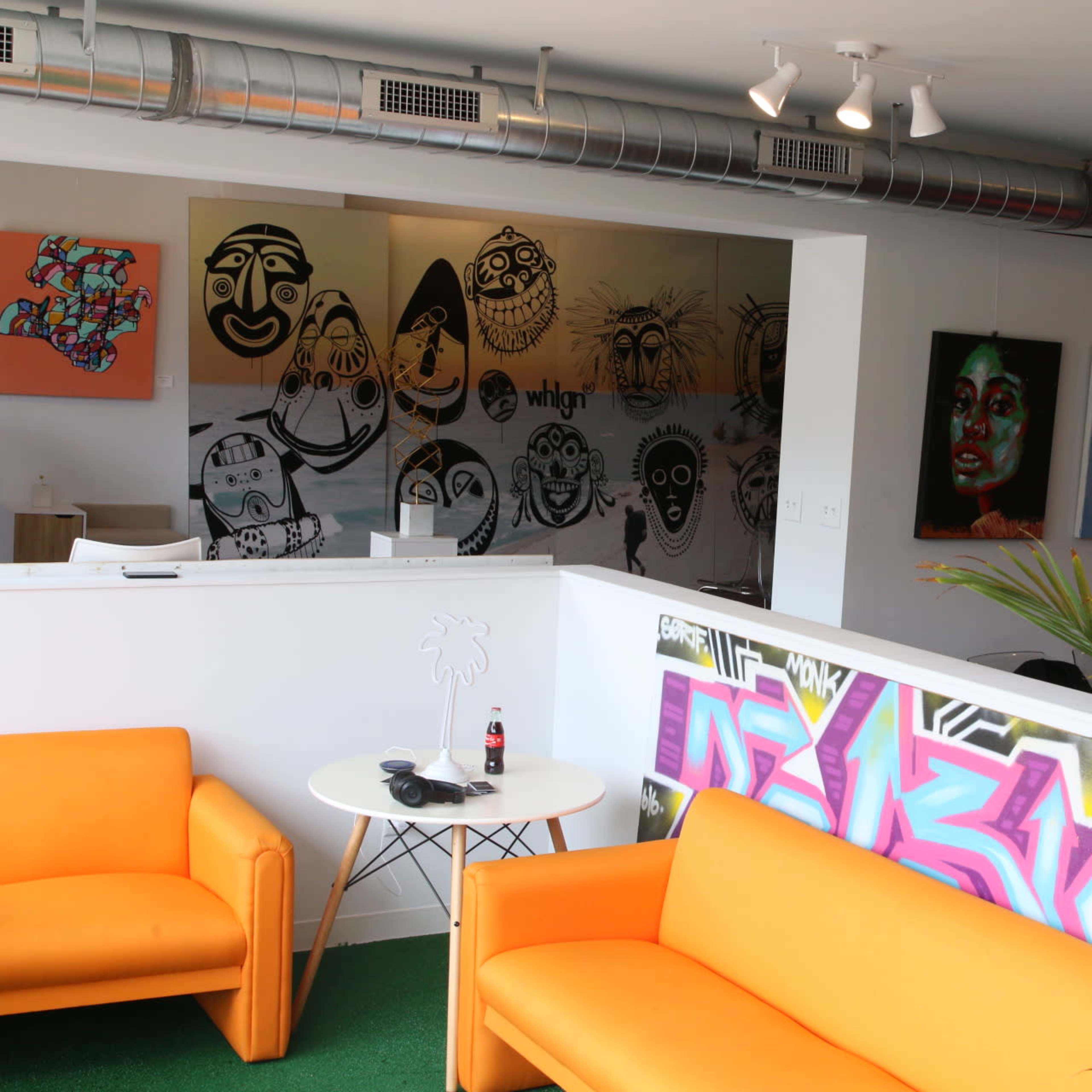 A modern office space with two orange sofas, a white partition featuring graffiti art, and several colorful paintings of faces on the walls.