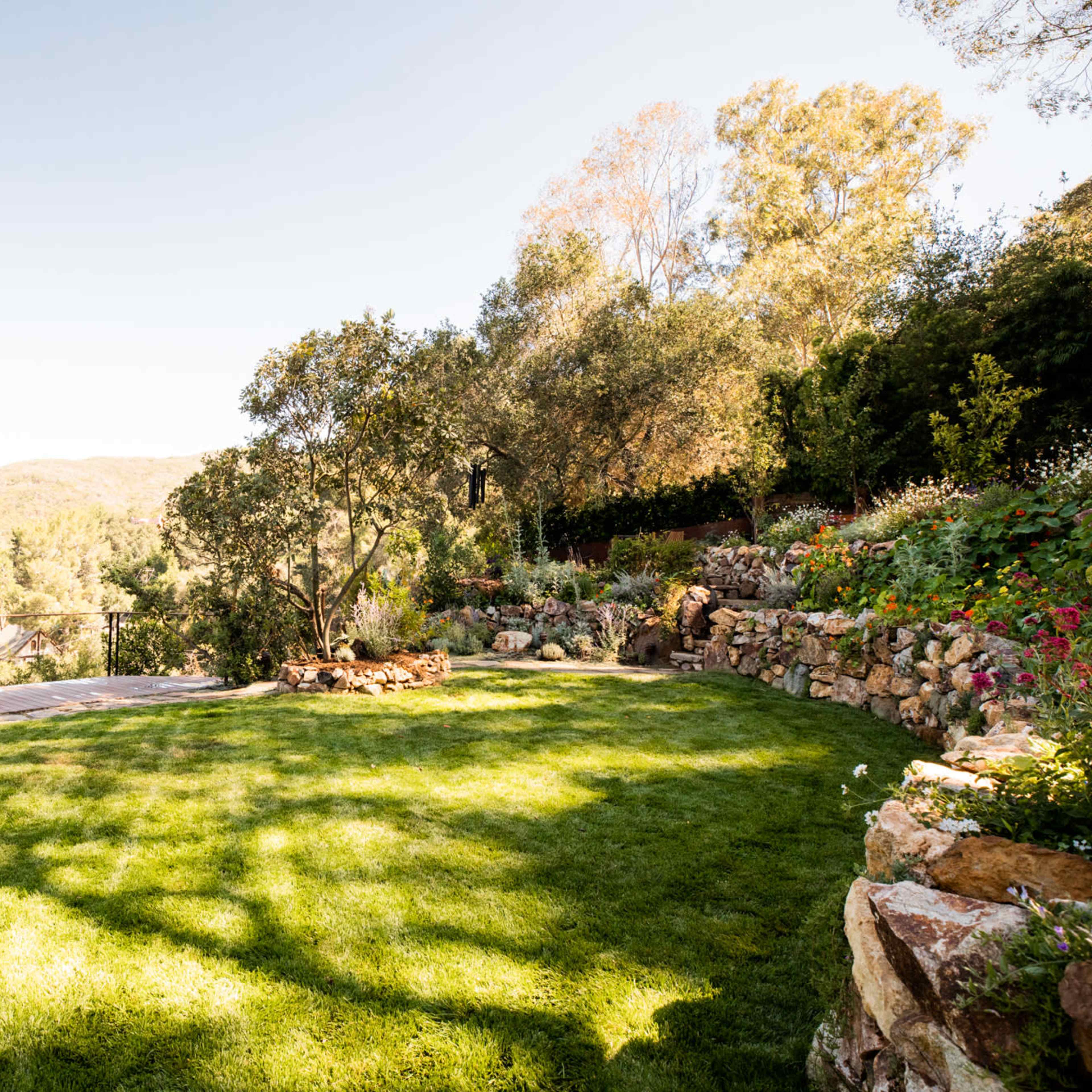 A well-maintained grassy area is surrounded by trees and a stone wall, with a wooden deck in the background and flower beds on the right.