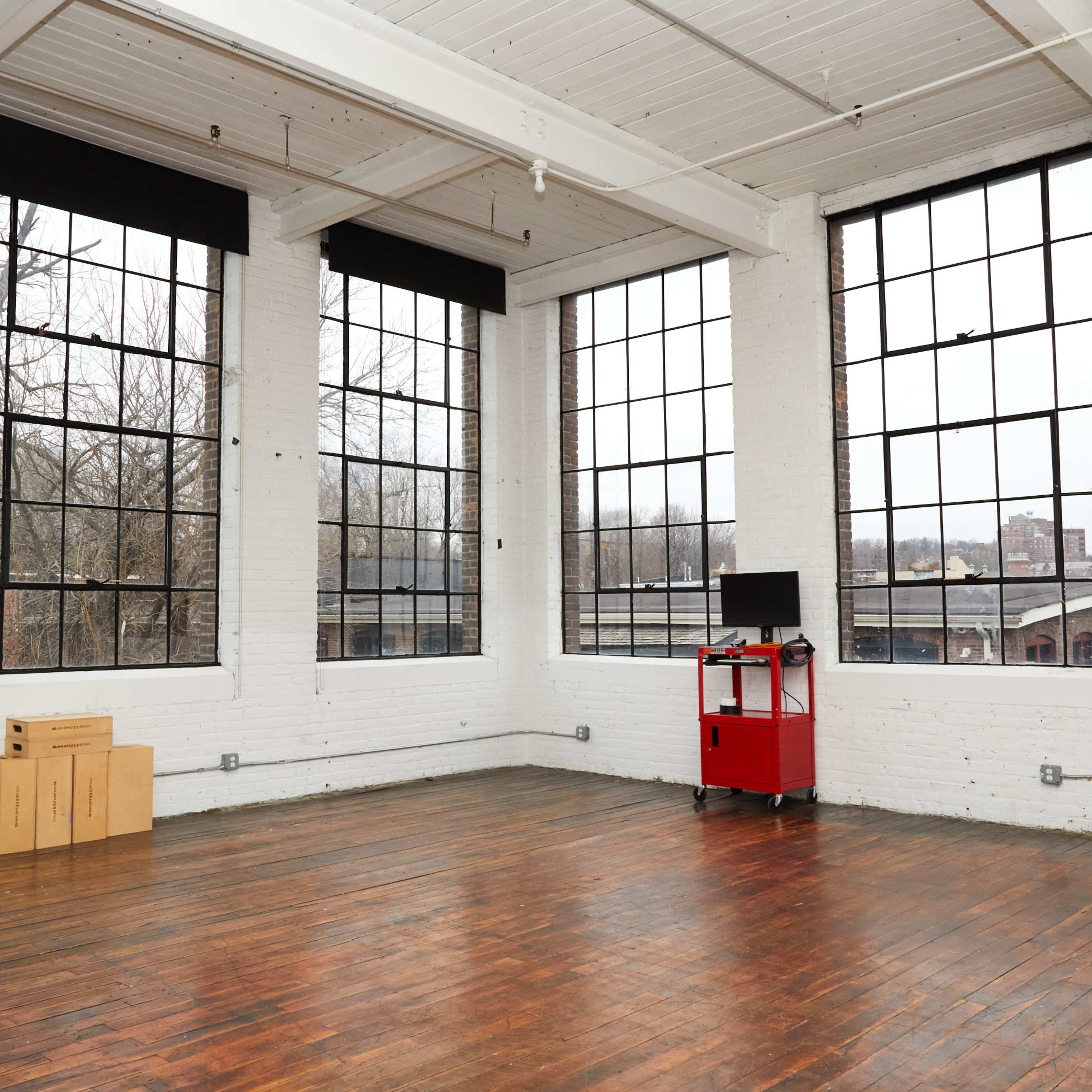 A spacious room with large windows, wooden floors, a red cart, and a mirror against a white brick wall.