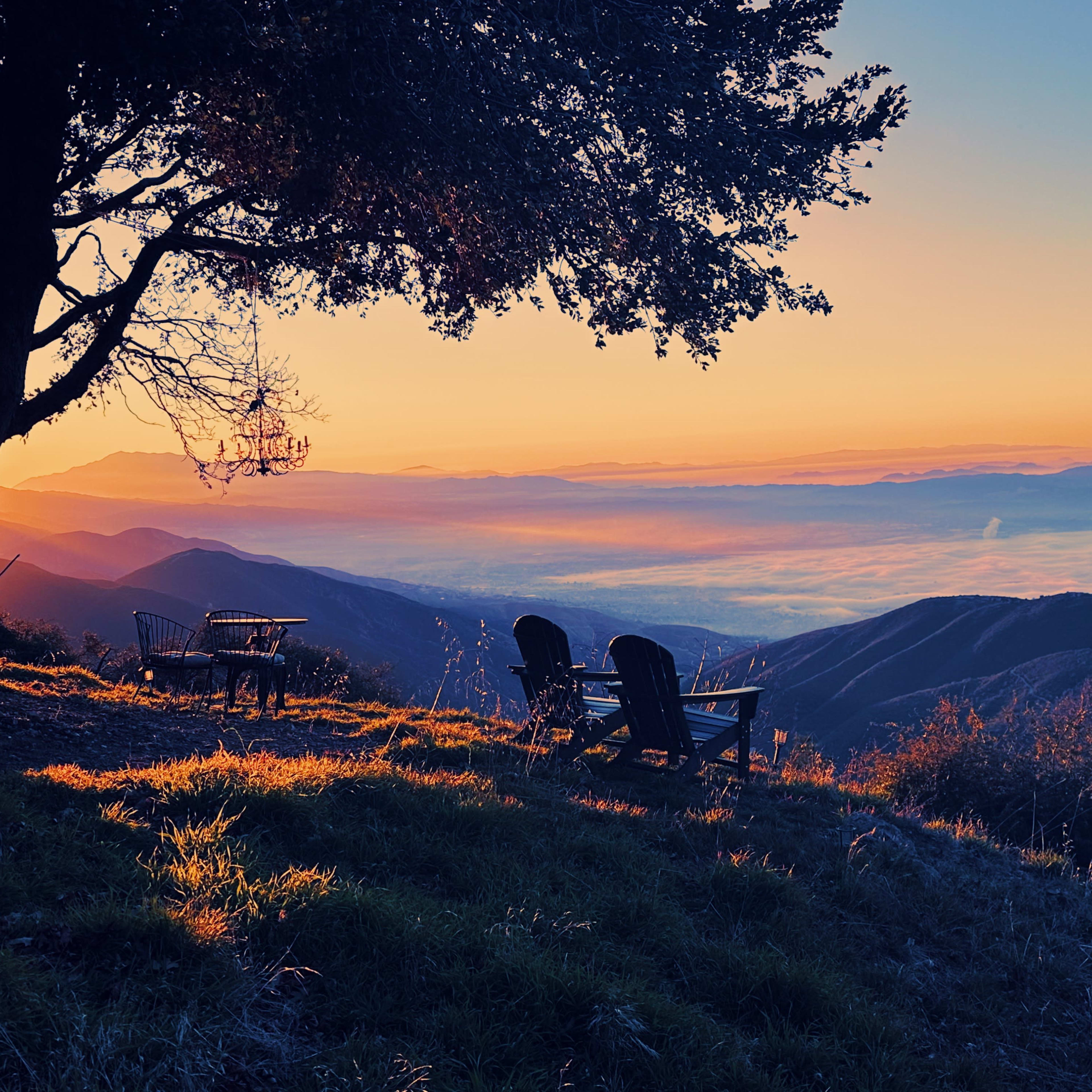 Two wooden chairs sit beside a tree overlooking a valley at sunset, with mist rising from the landscape below.
