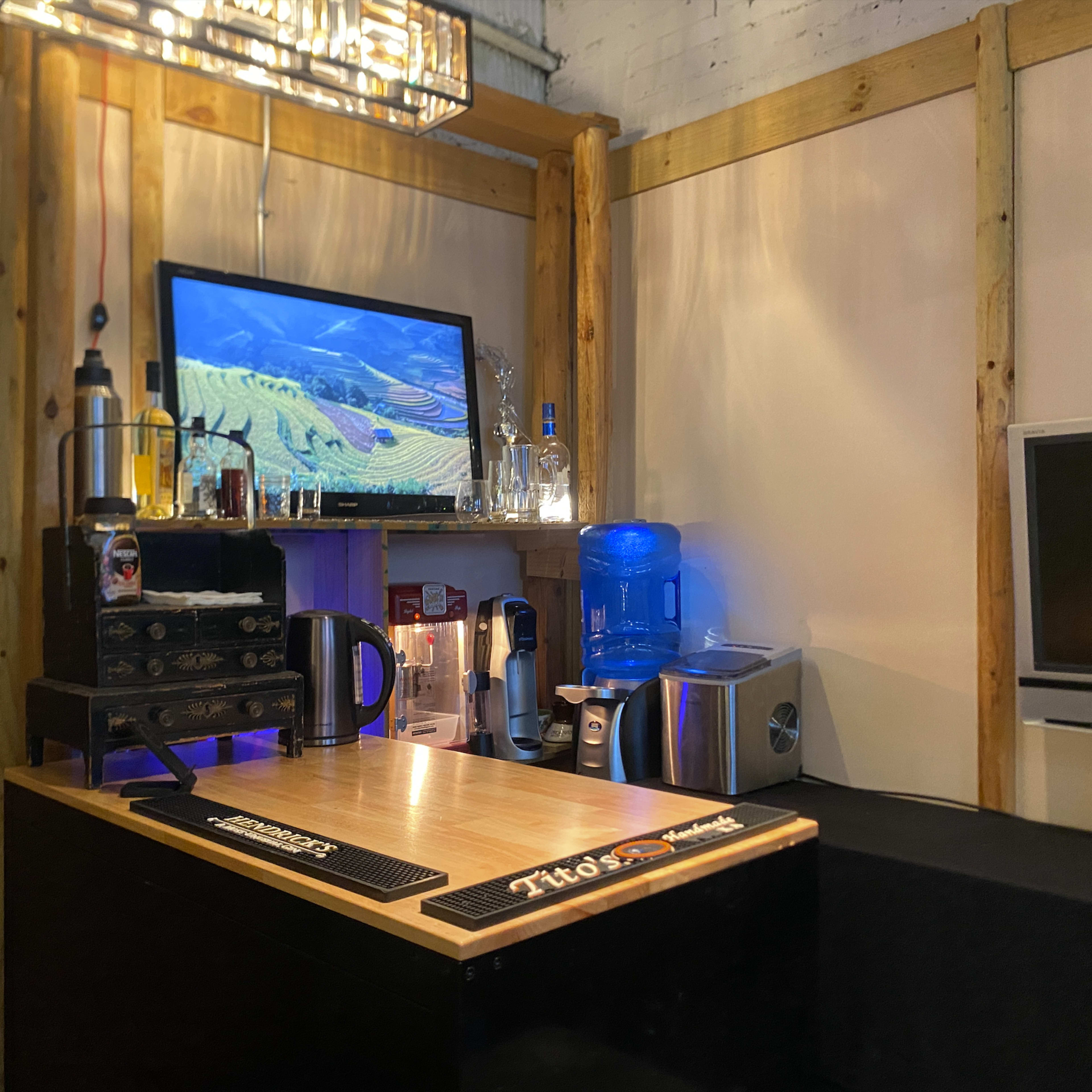 A bar area equipped with a wooden countertop, a television displaying a landscape, bottles of alcohol, a coffee maker, and a water cooler.