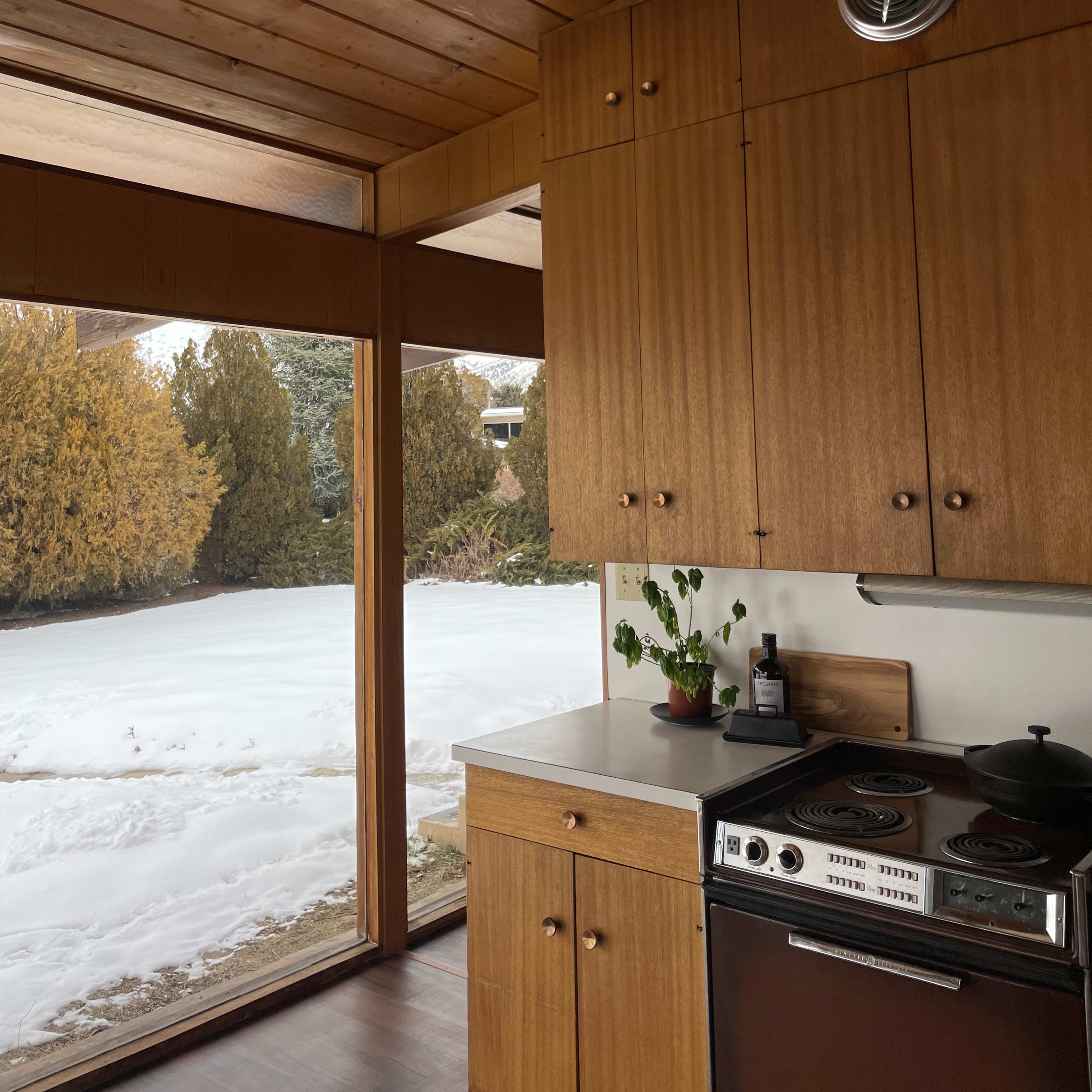 A wooden kitchen with a vintage stove and a view of a snow-covered garden through large glass windows.