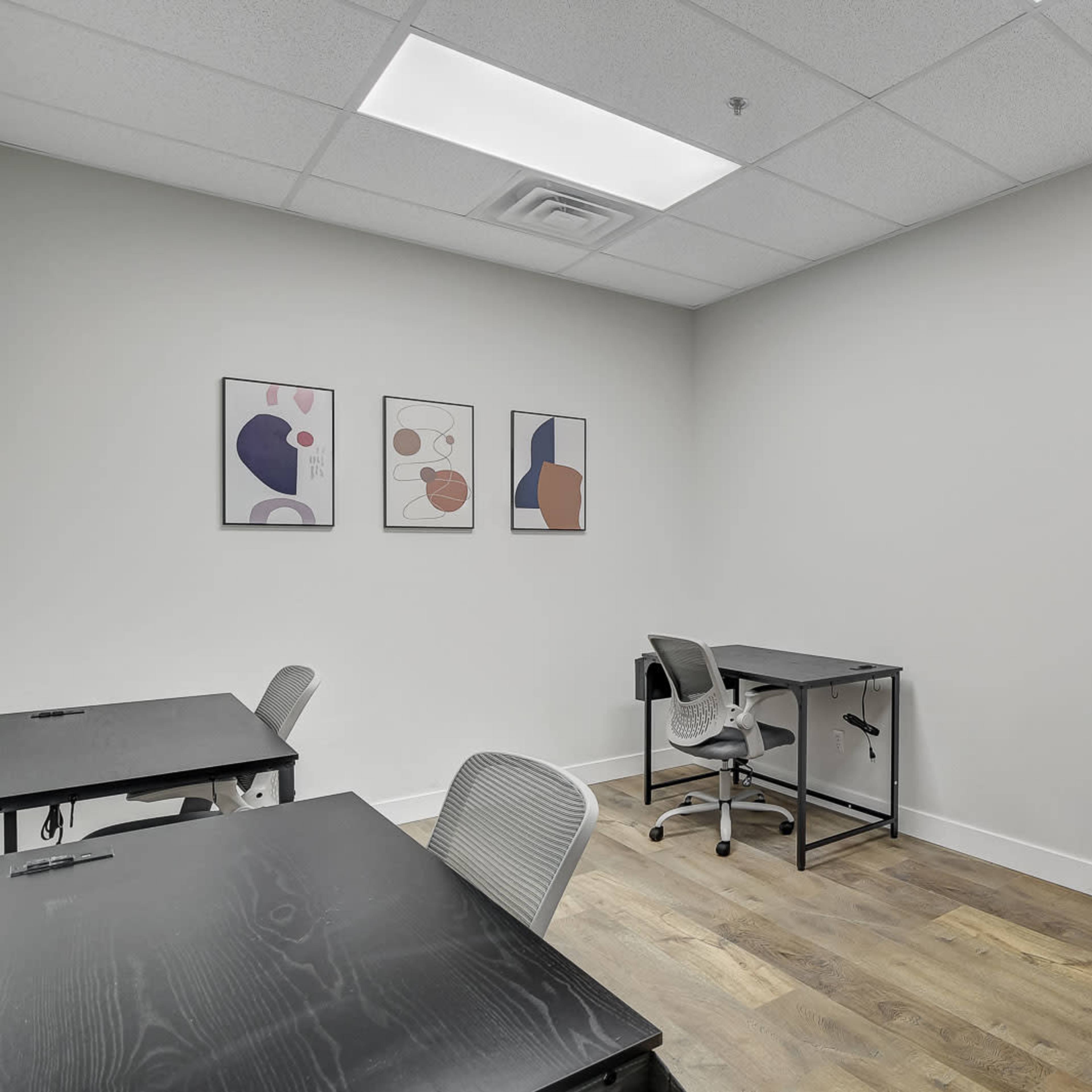 A minimalistic office space with two black desks, a chair, and three abstract art pieces on the wall.