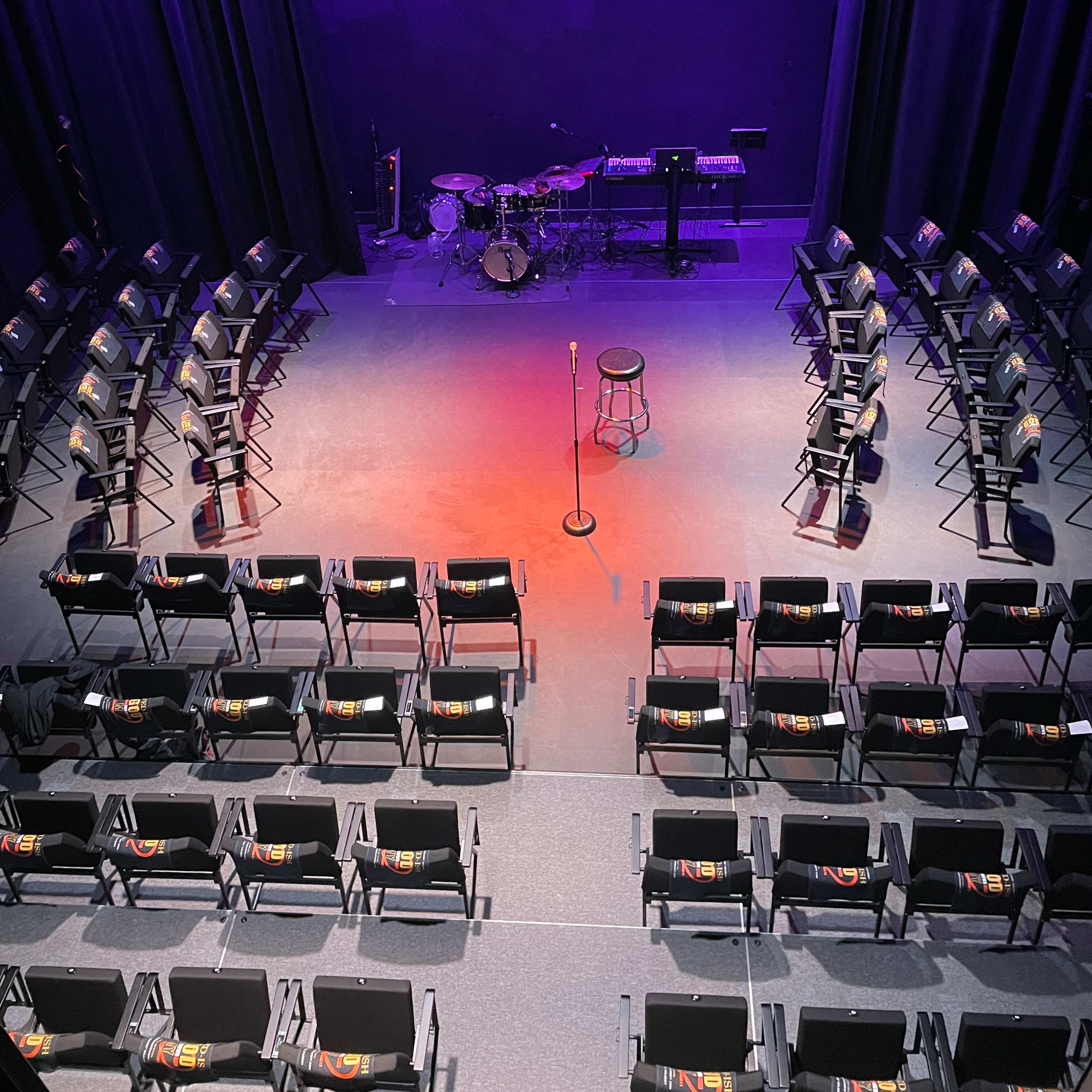 A stage set up for a performance, featuring a microphone on a stand, a stool, and a drum set, surrounded by rows of empty chairs.
