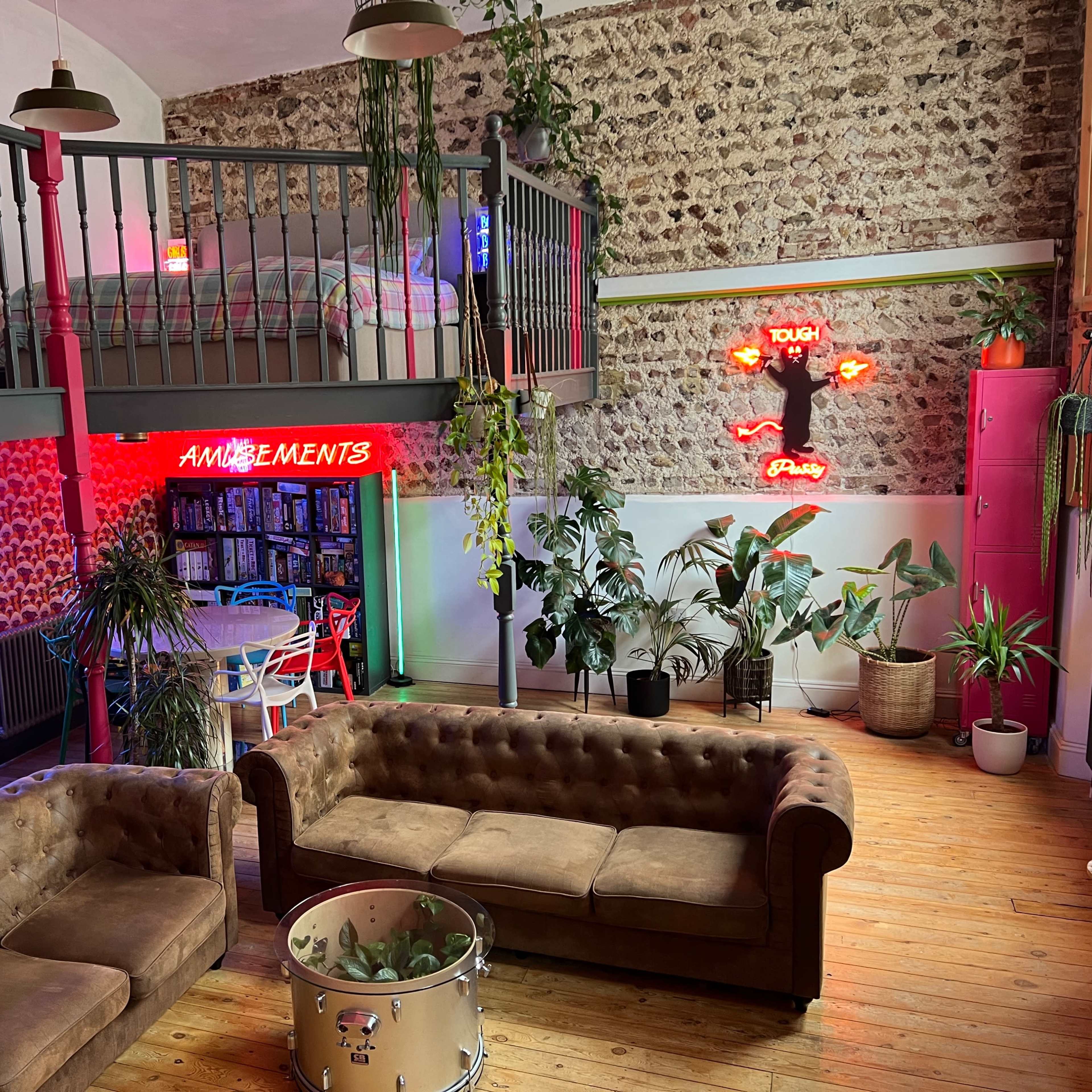 A spacious room featuring a loft area with a bed, two brown leather sofas, vibrant neon signs, and various indoor plants.