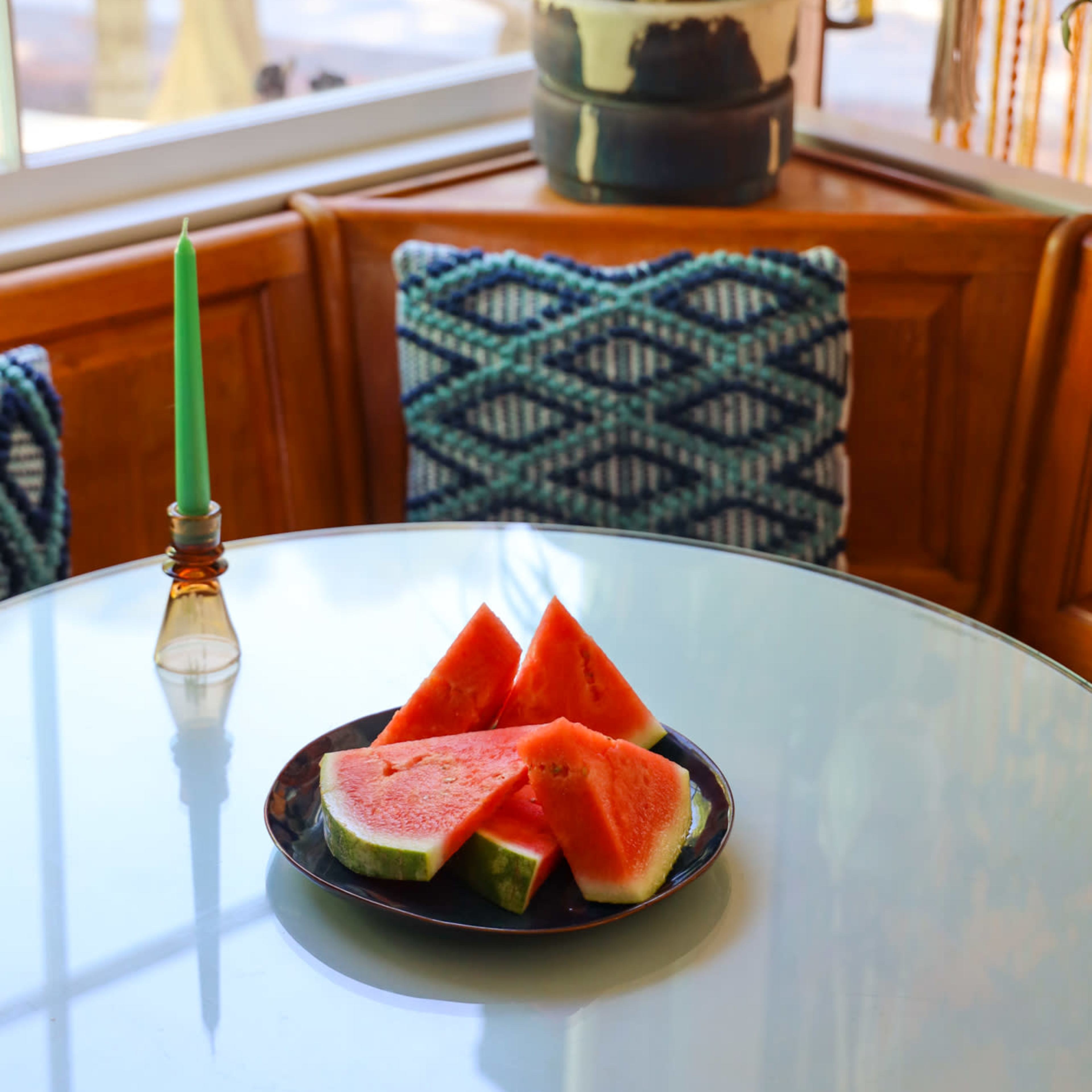 A plate of watermelon slices sits on a glass table beside a green candle in a decorative holder and patterned cushions.