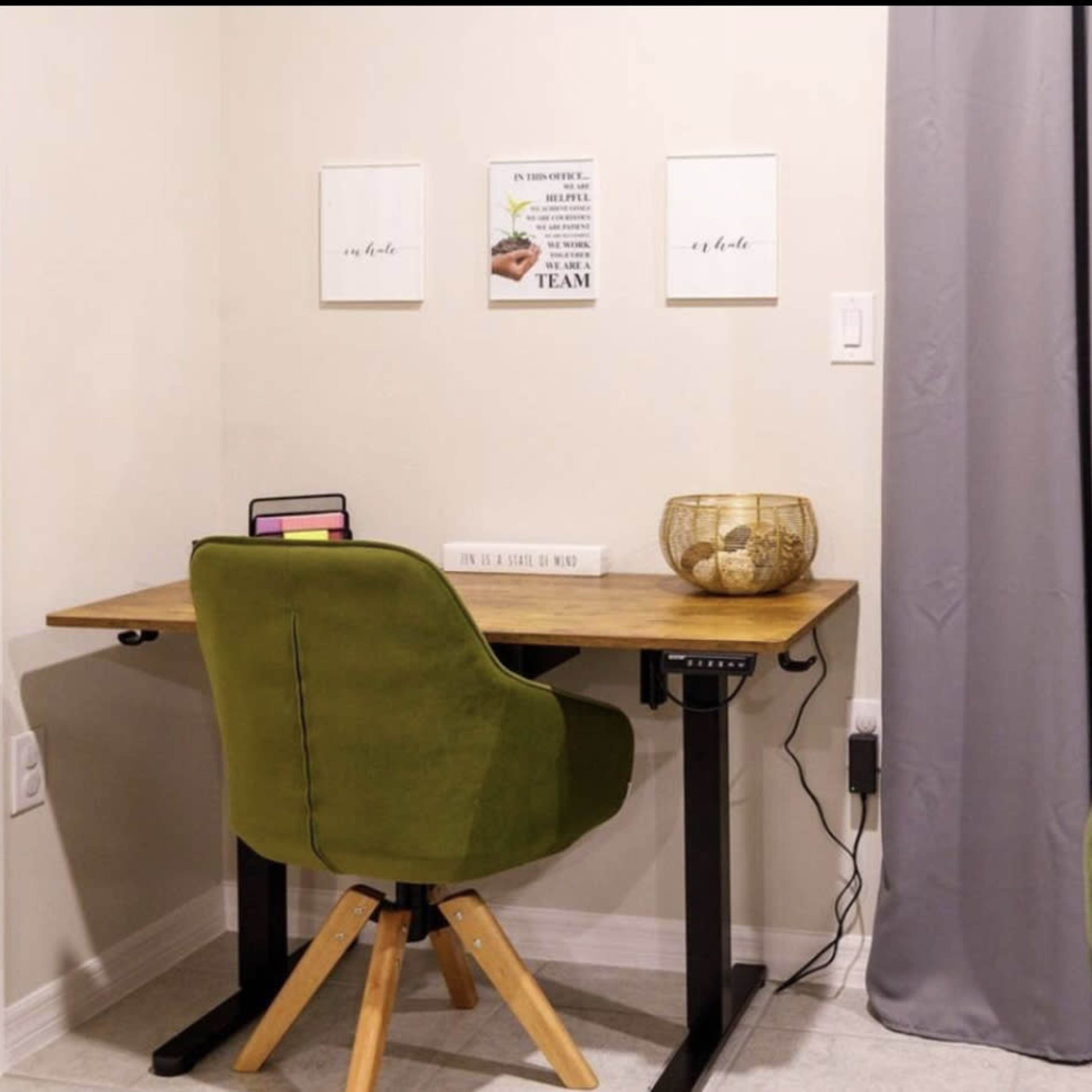 A wooden desk with a green chair is positioned in a corner of a room, accompanied by framed wall art and a decorative bowl on the desk.