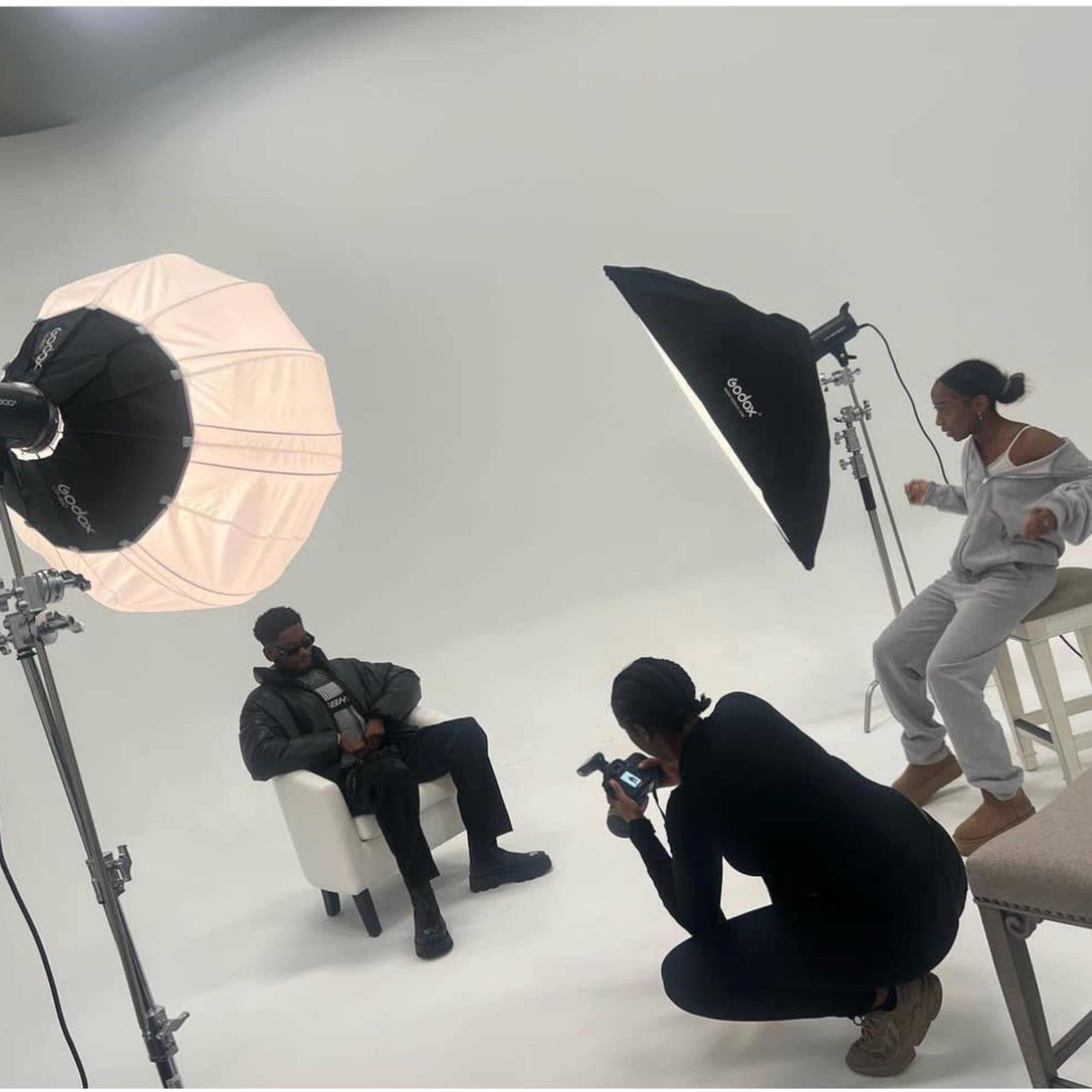 A photographer is capturing an image of a man sitting in a chair, while a second person adjusts the lighting in a studio setting.