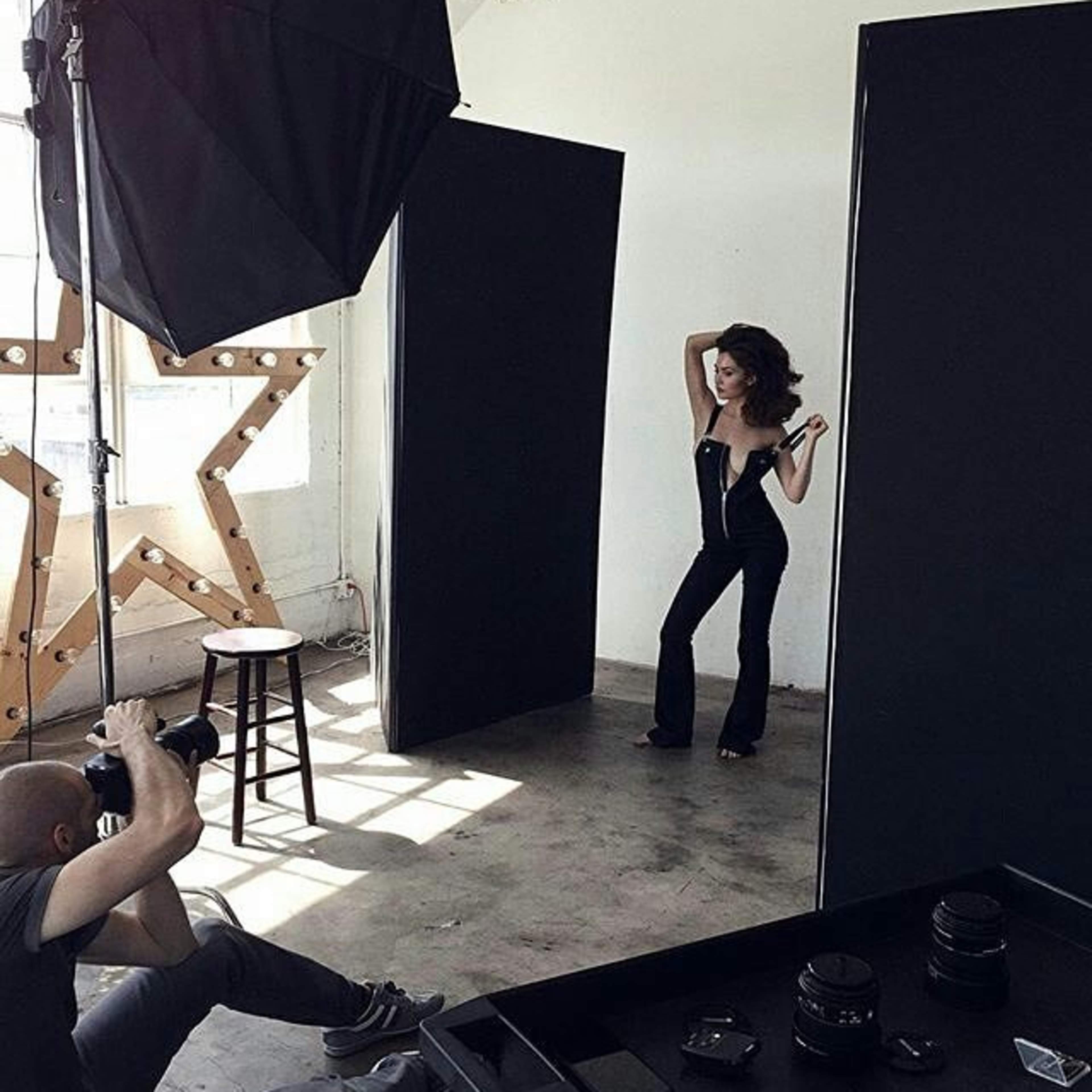 A photographer captures a model posing in a studio surrounded by large black backdrops and soft lighting equipment.