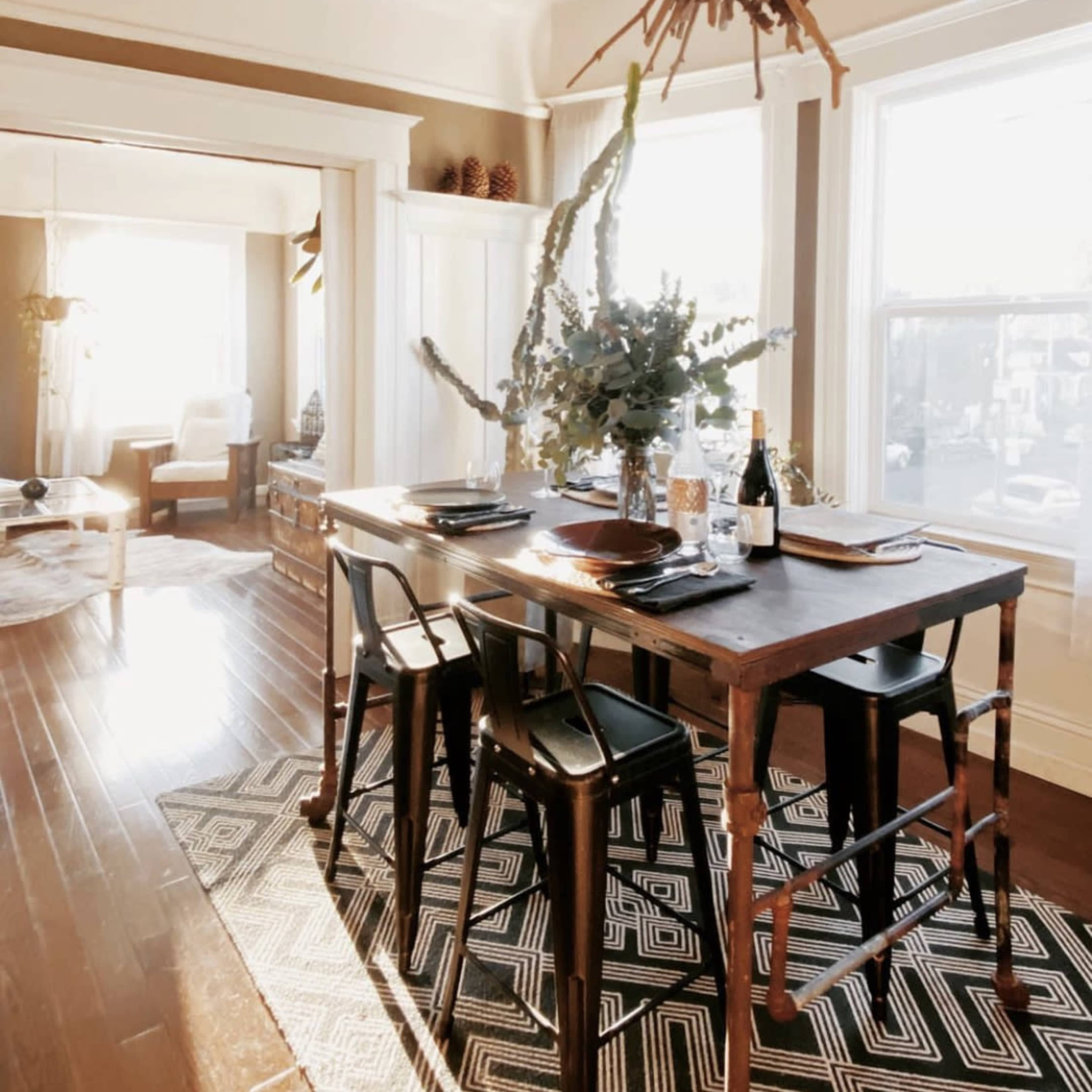 A wooden dining table set for a meal is positioned in a brightly lit room with large windows, featuring a decorative centerpiece made of branches and greenery.