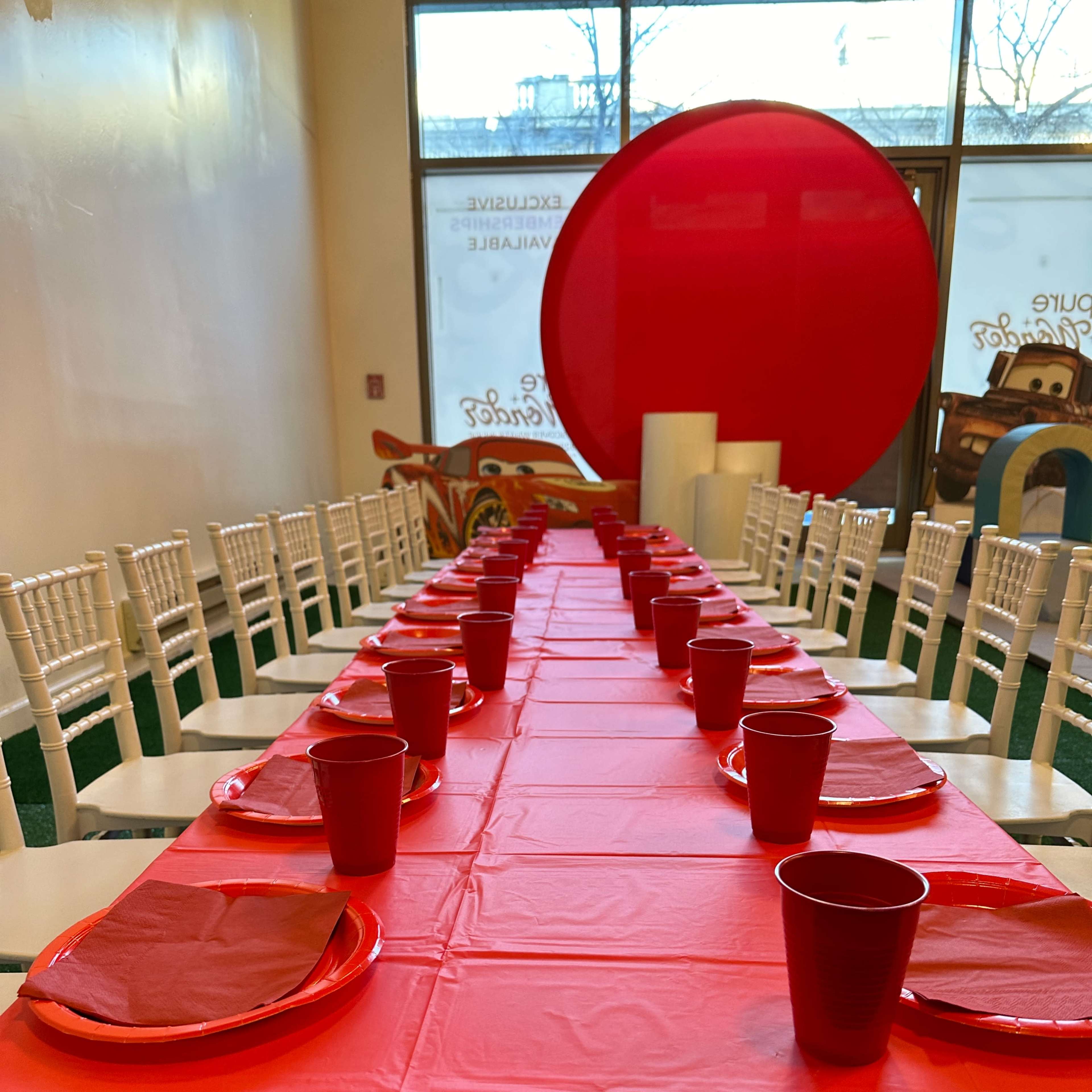 A long table is set with red tablecloths, plates, and cups in a brightly lit room with large windows.