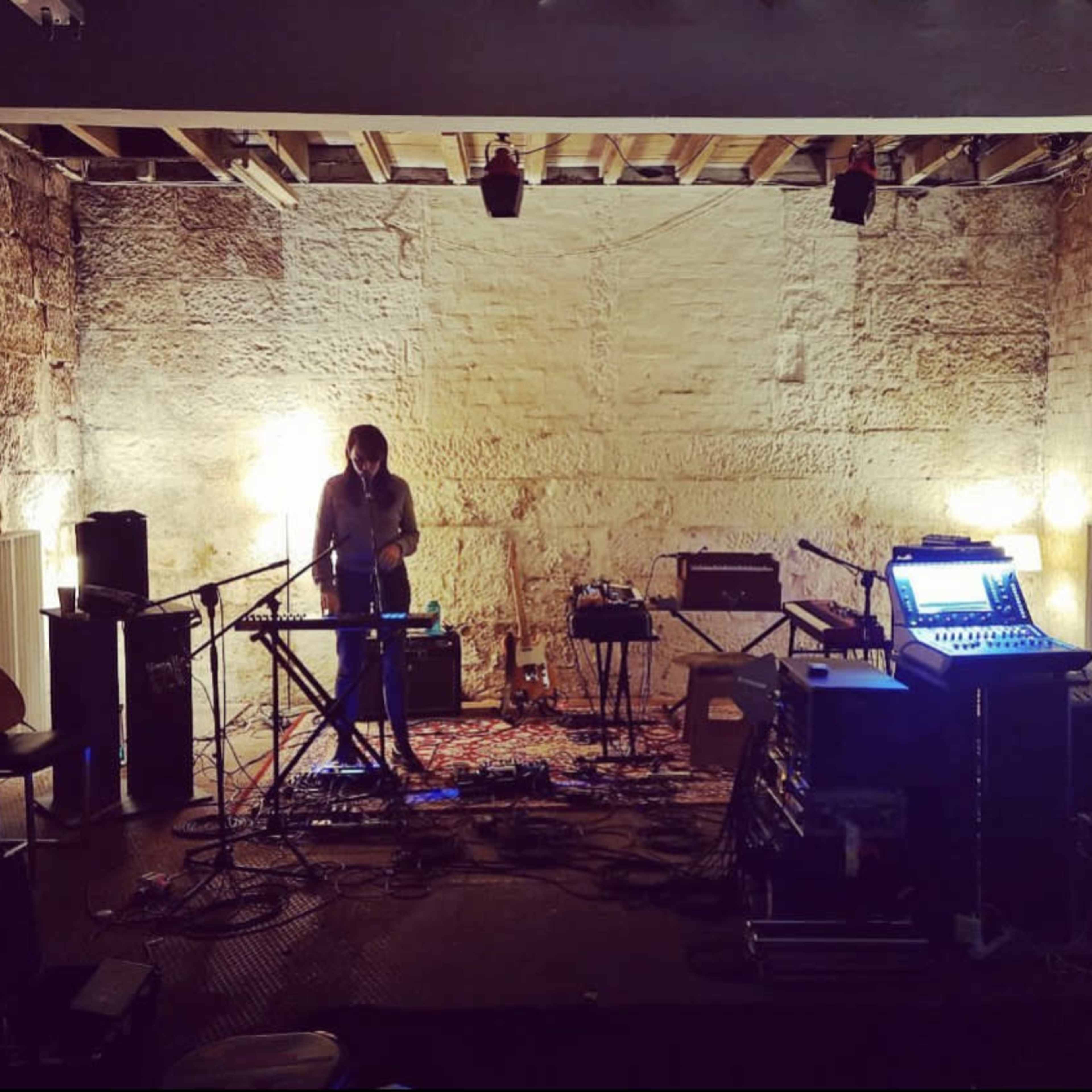 A musician stands at a keyboard in a sparsely furnished recording studio with exposed stone walls and various instruments and audio equipment arranged around the space.