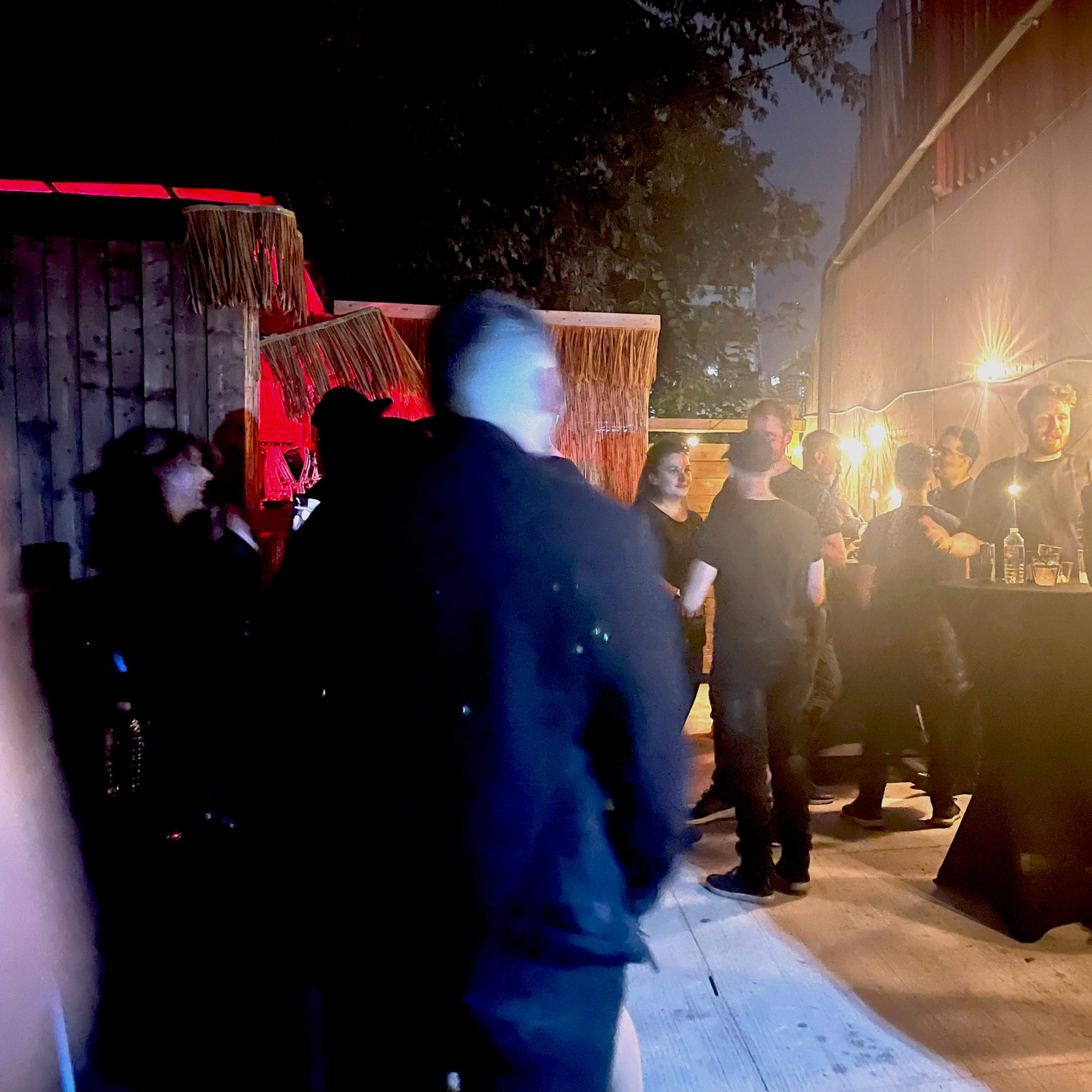 A group of people gather at a dimly lit outdoor venue with string lights and a bar table.