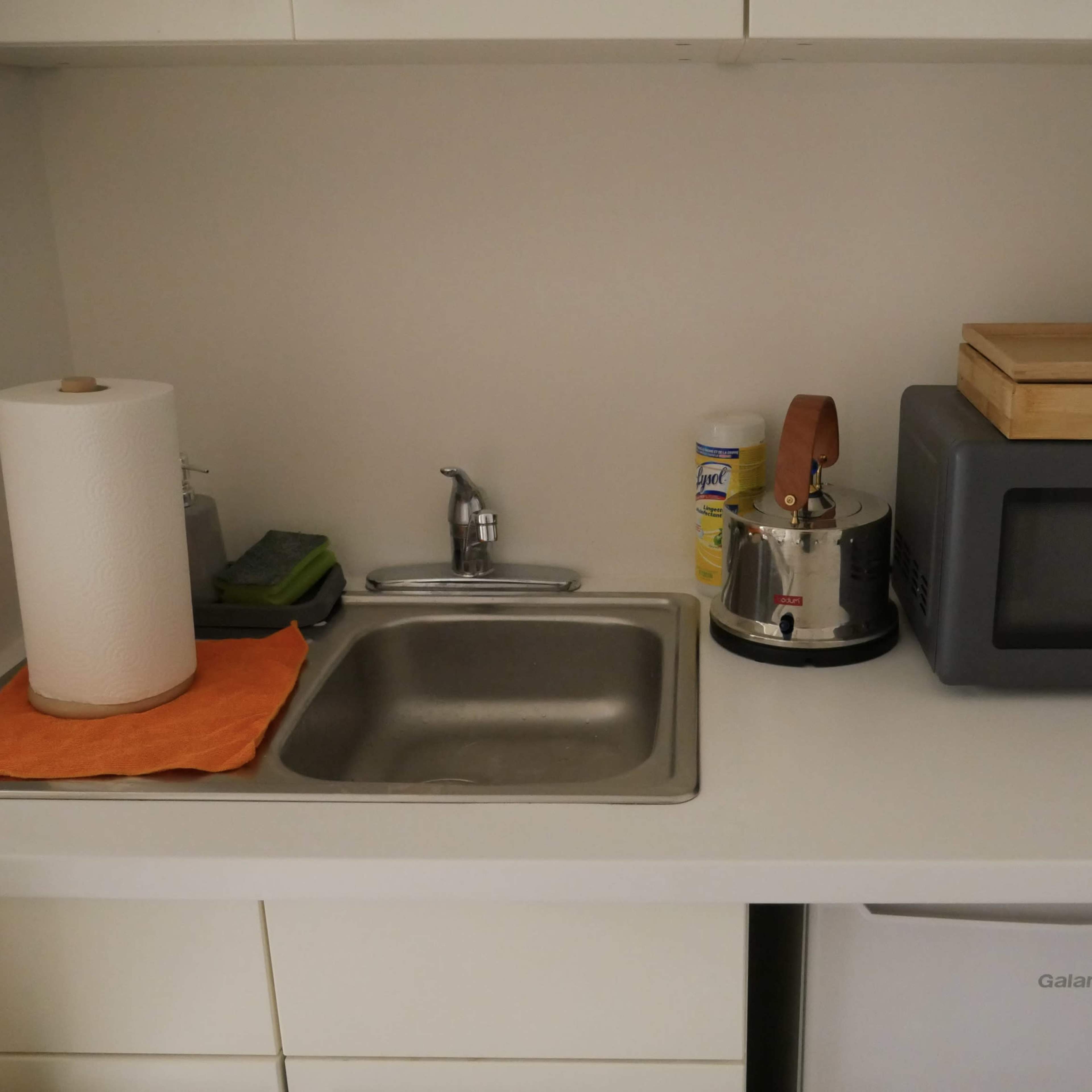 A kitchen countertop with a sink, a microwave, a kettle, a paper towel roll, and a tissue box.