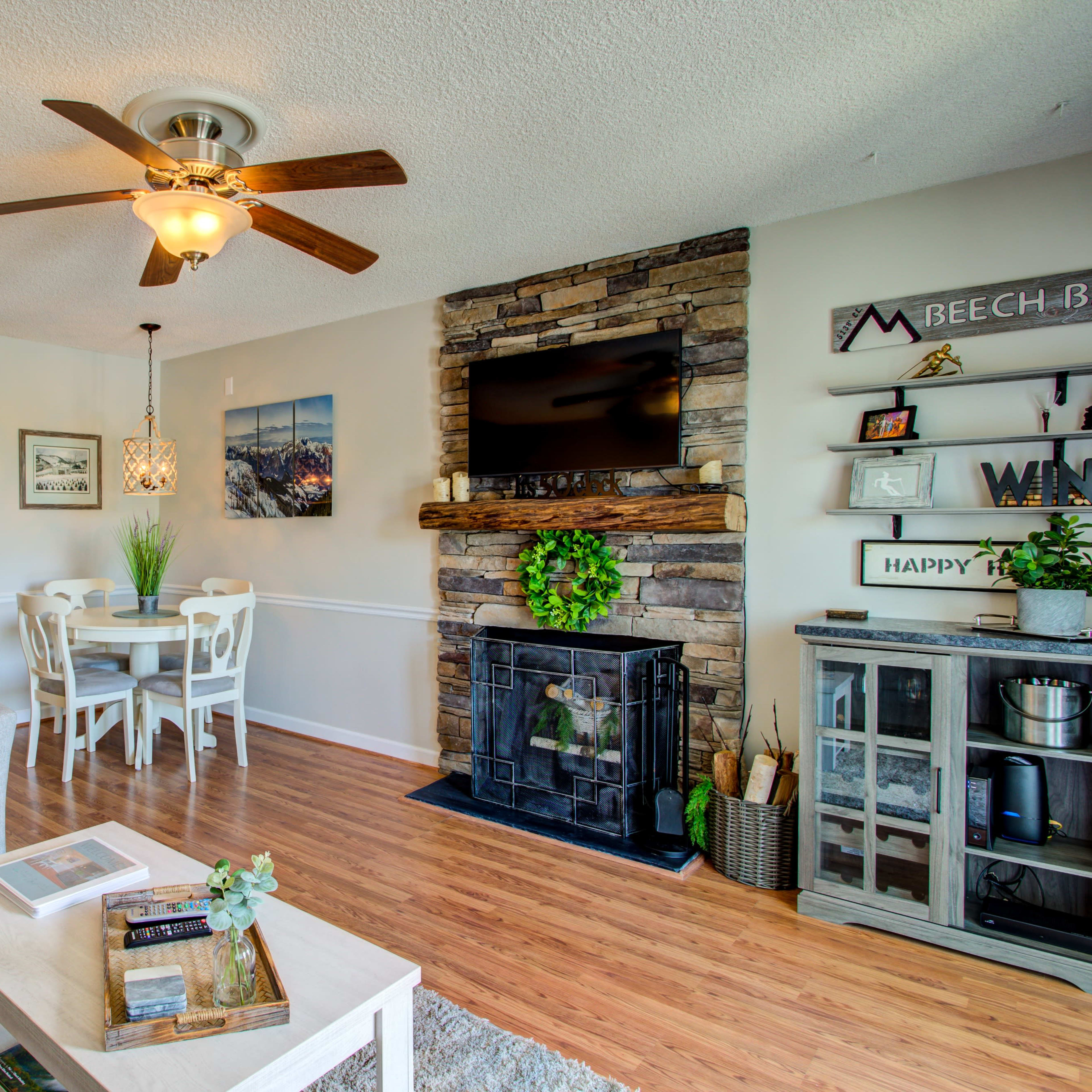 A living room with a stone fireplace, a ceiling fan, a dining area, and modern furnishings.