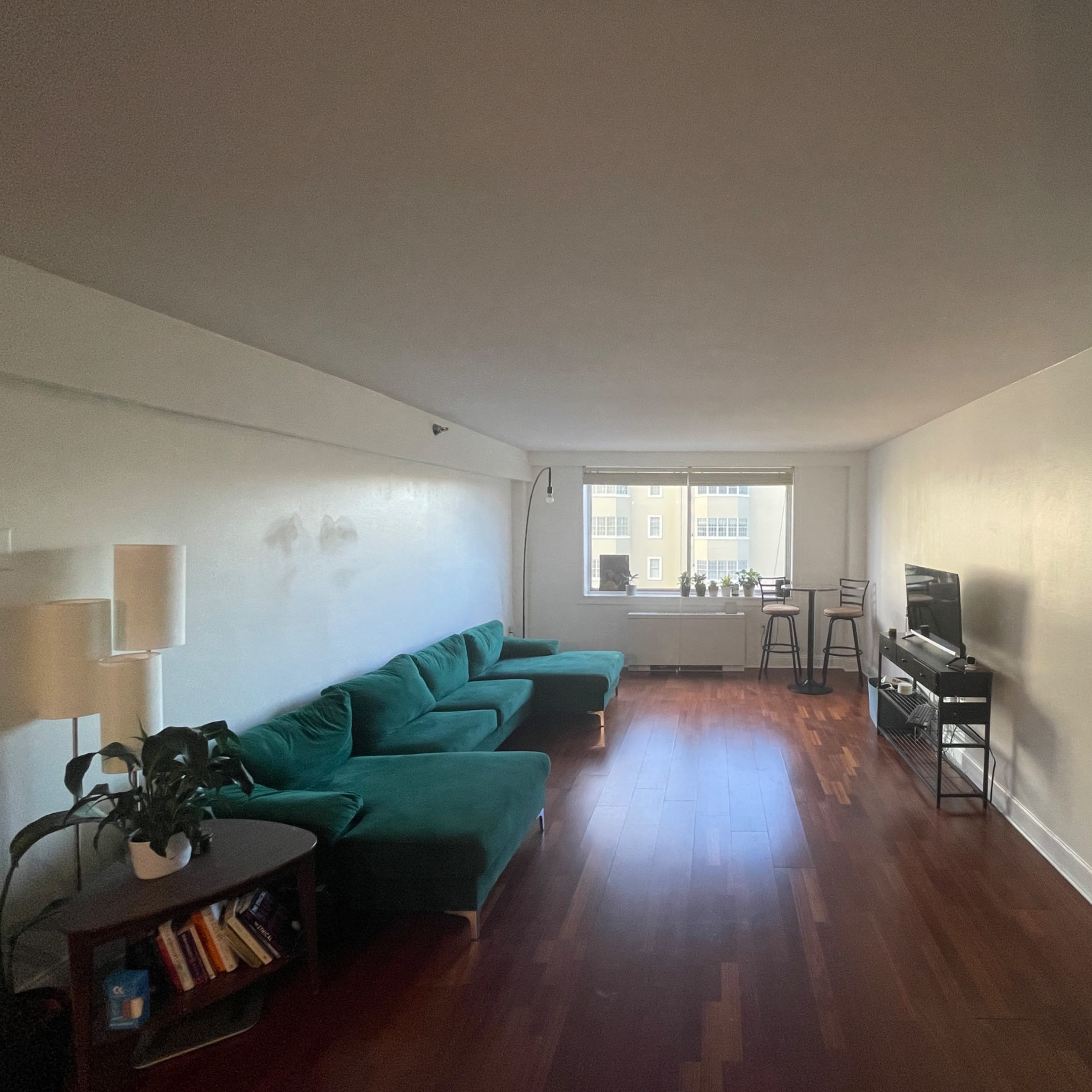 A spacious living room with a green sectional sofa, a small table with books, a TV stand, and a window overlooking a building across the street.