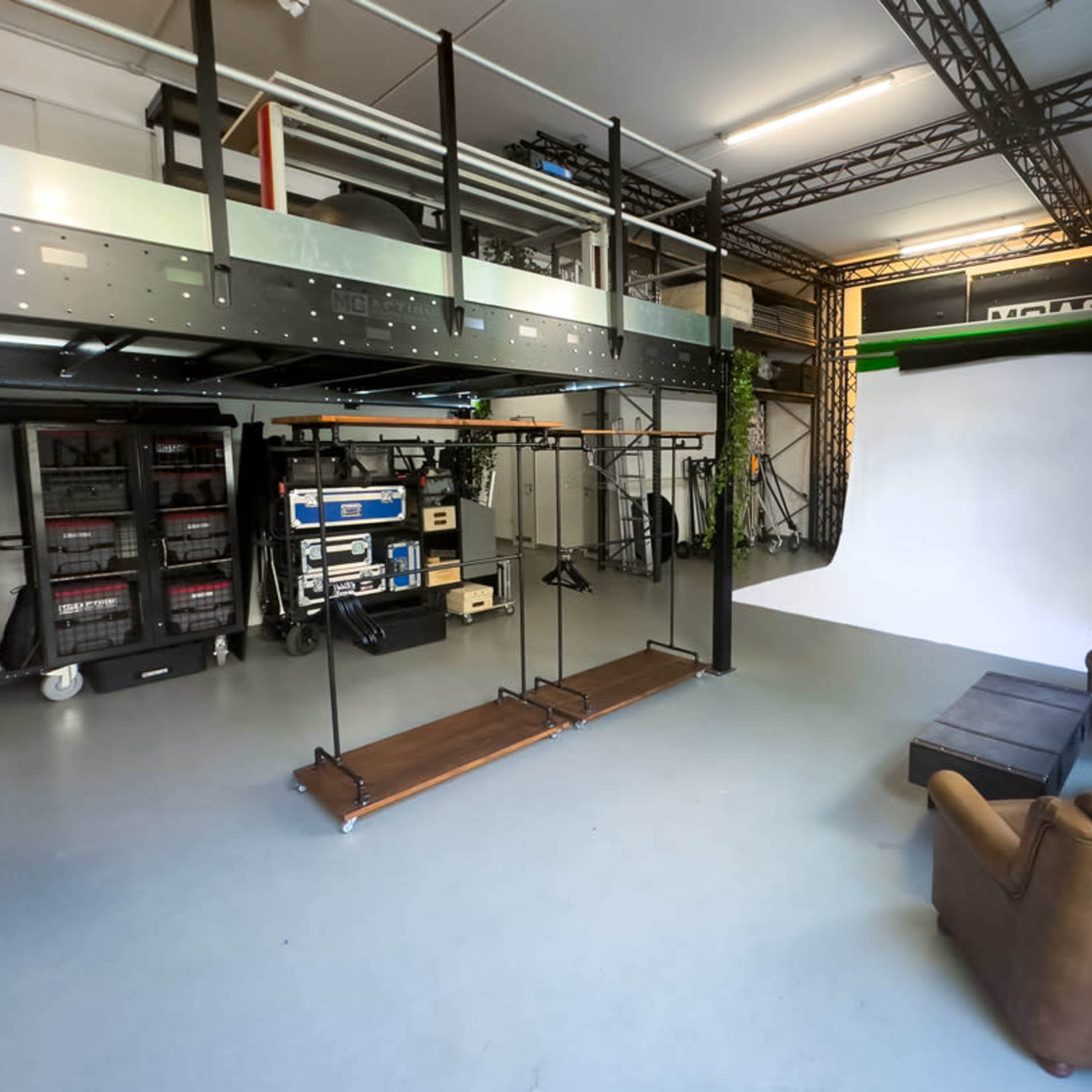 A spacious studio environment featuring a white backdrop, a seating area with brown chairs, and various technical equipment organized on shelves.