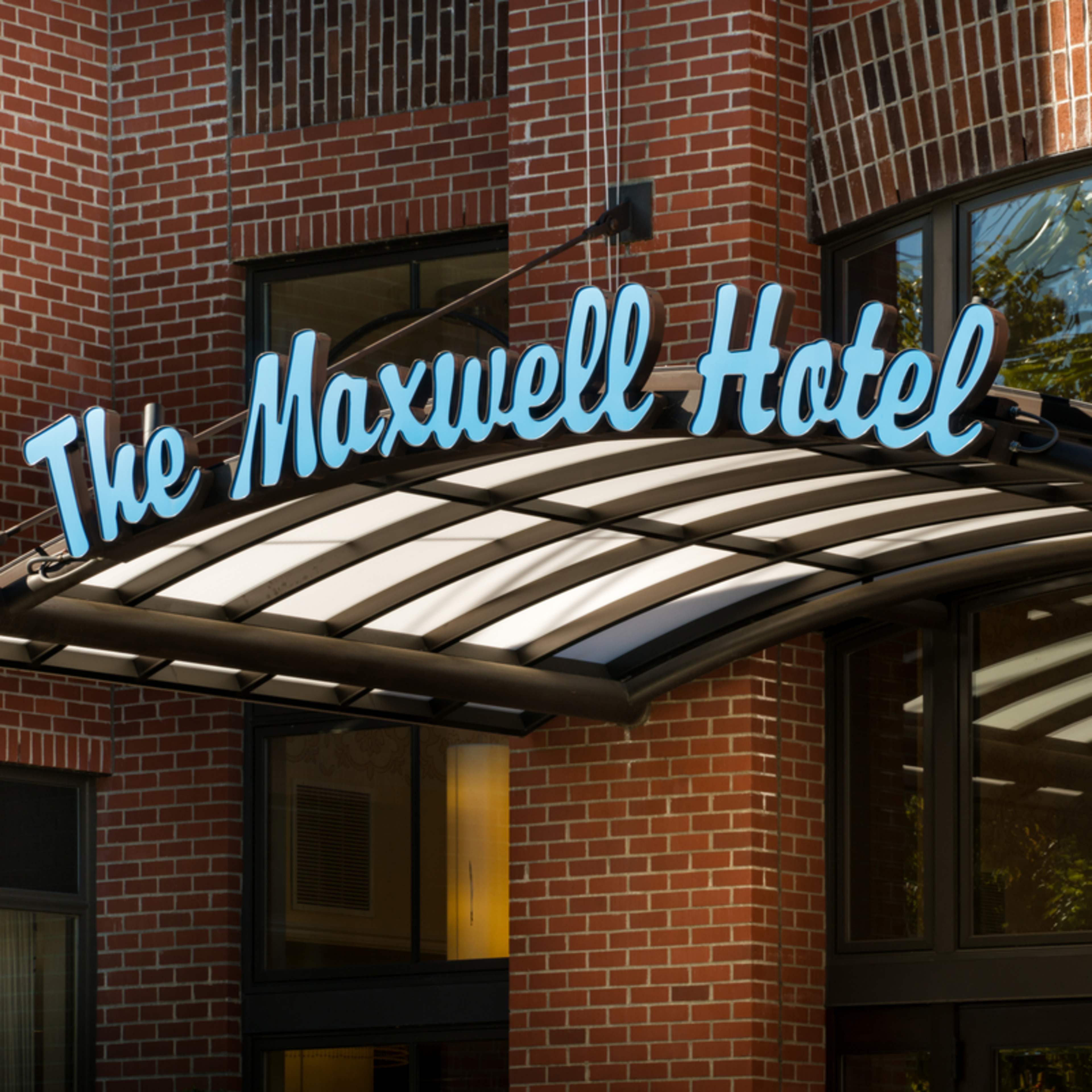 The exterior sign of The Maxwell Hotel, featuring a blue, illuminated name mounted above an entrance with a curved awning.