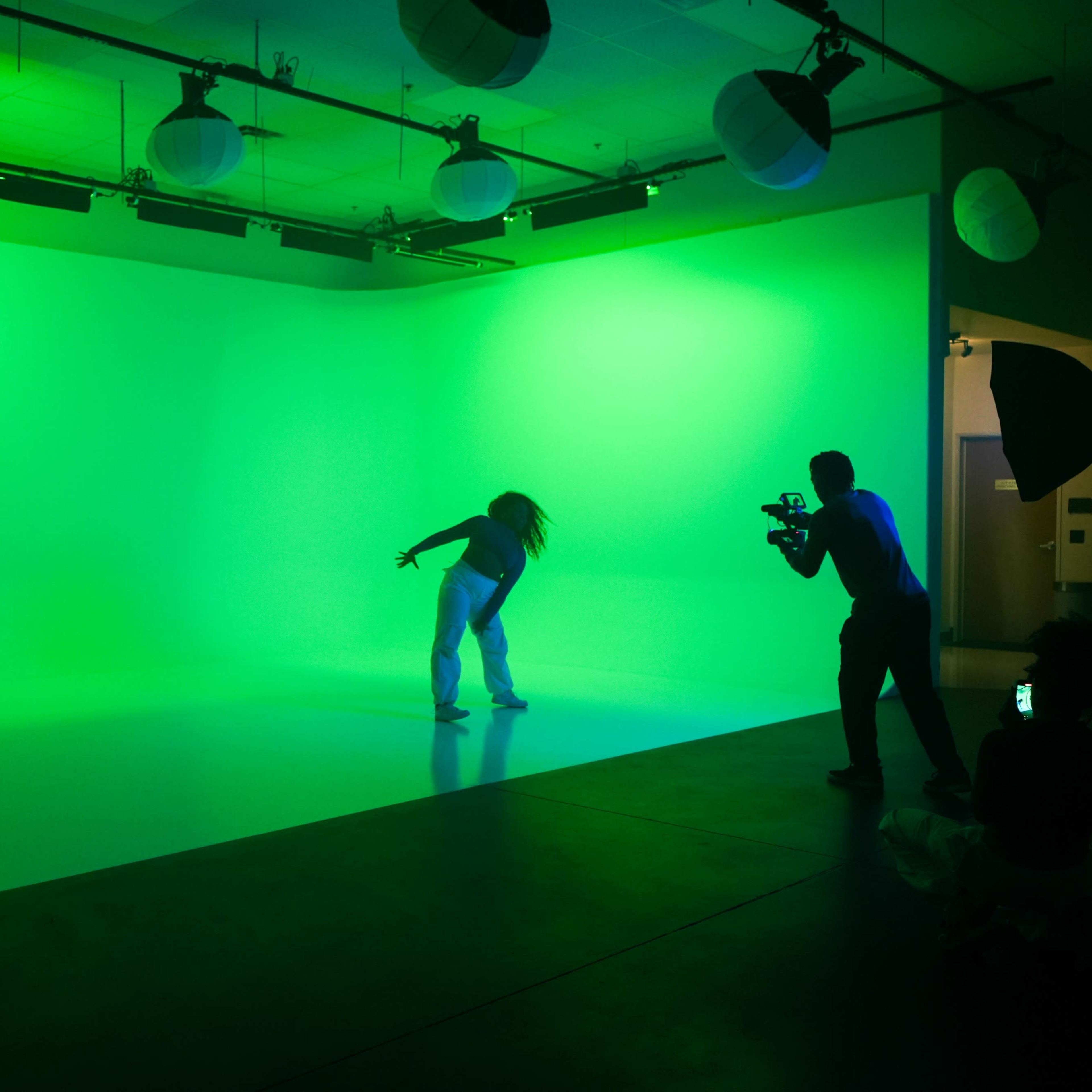 A person dances in a green-lit studio while another person records with a camera.