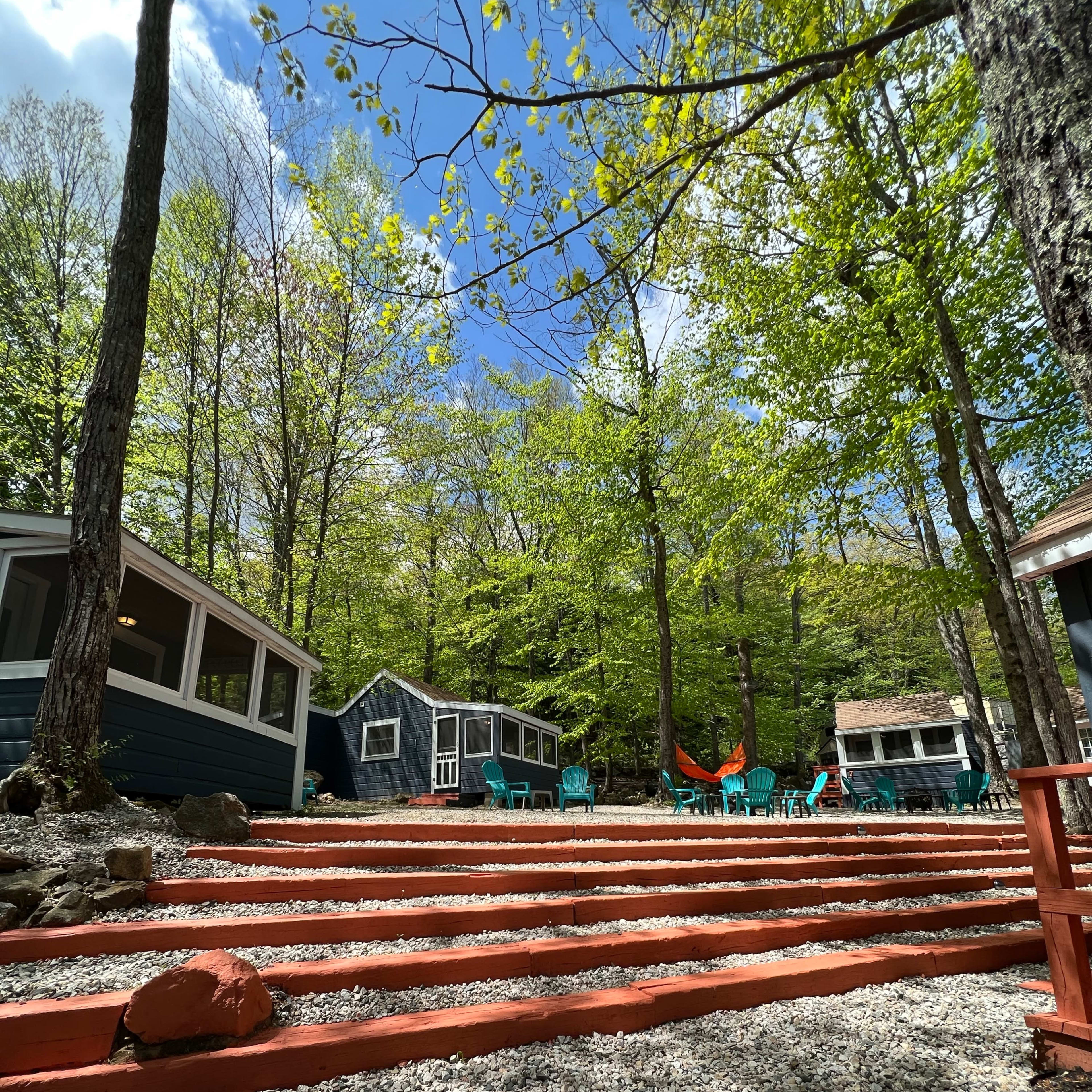 A wooded area with a series of cabins, stone steps, and turquoise chairs surrounded by trees under a clear blue sky.