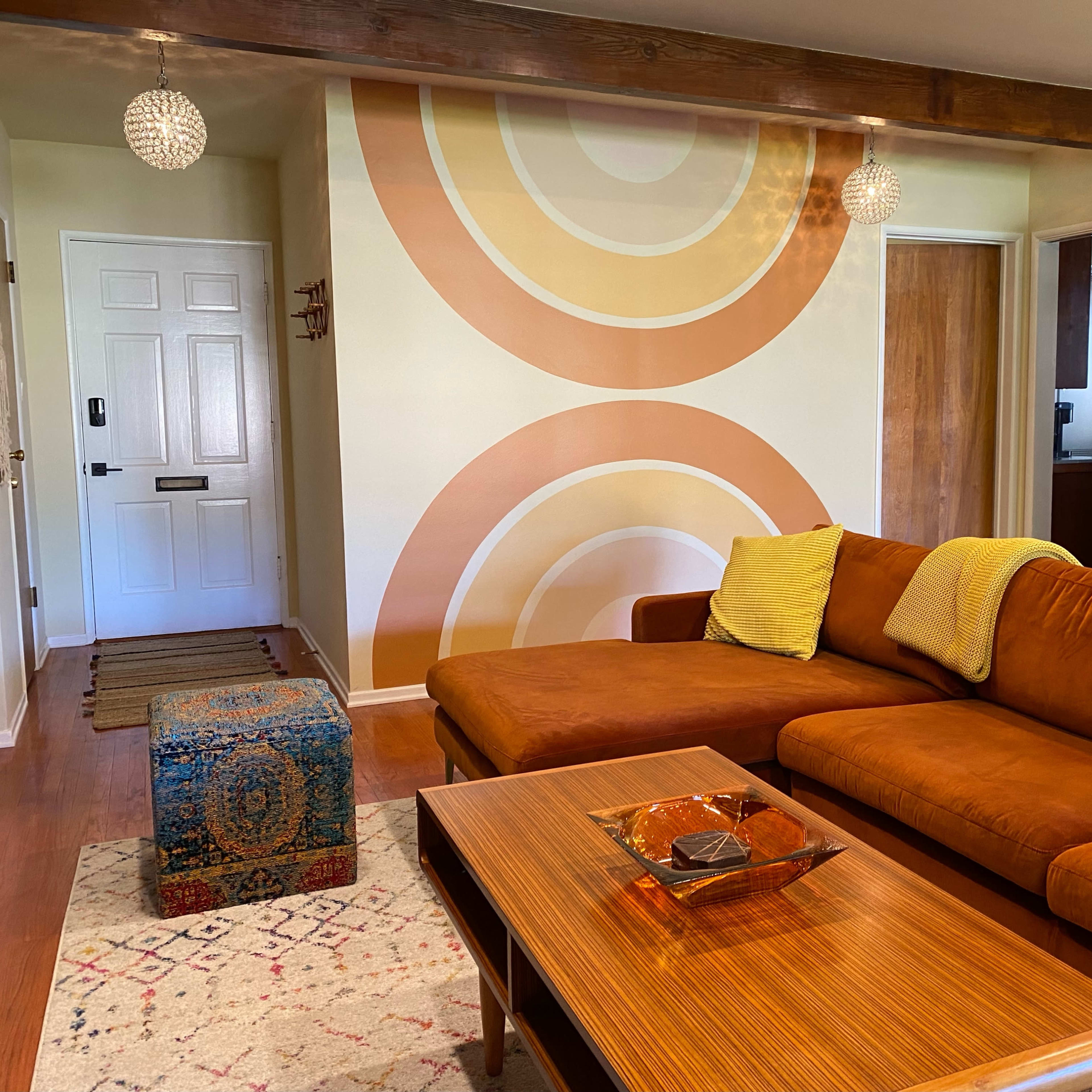 The living room features a brown sectional sofa with yellow throw pillows, a wooden coffee table, a patterned rug, and a retro wall mural of concentric circles.