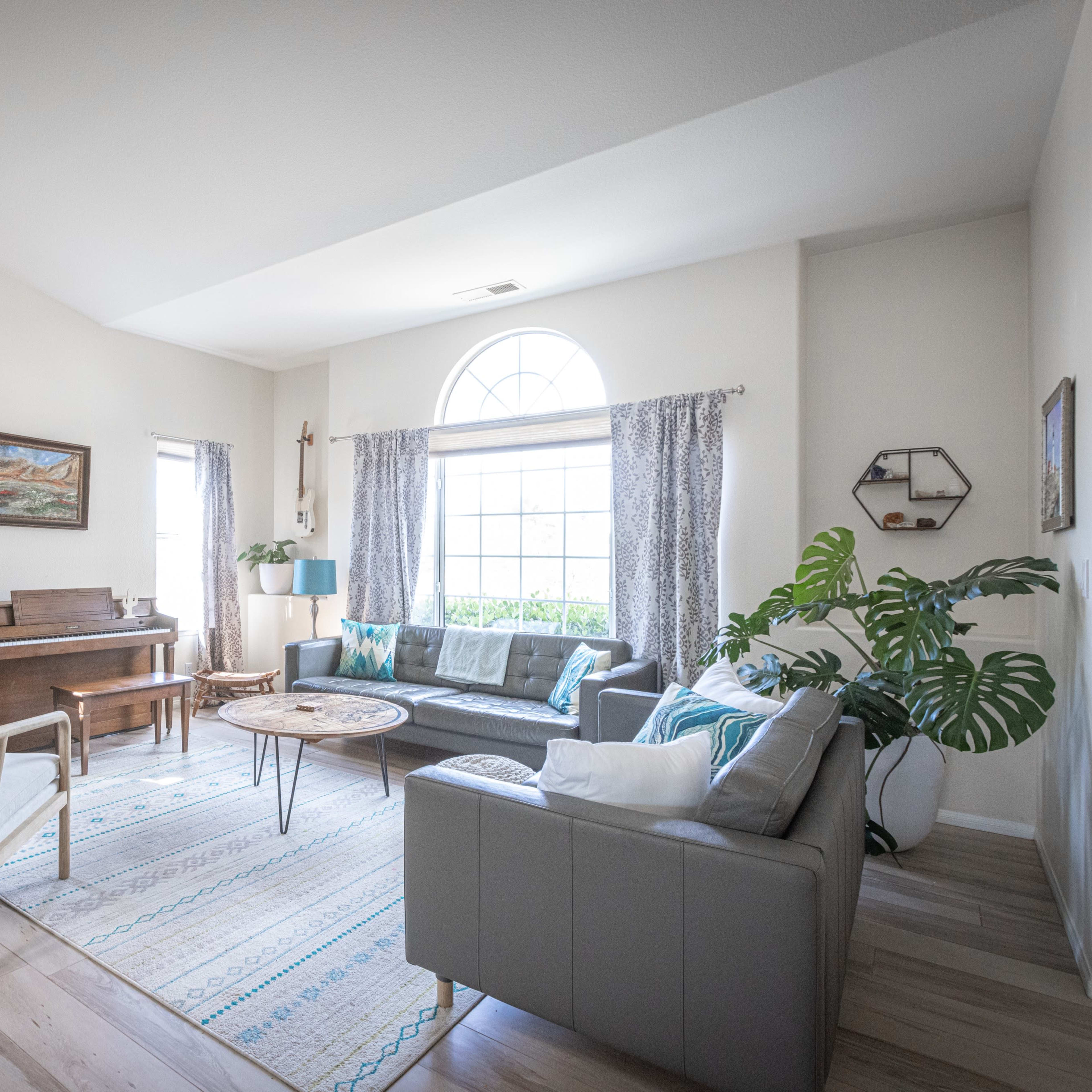 The living room features a piano, a light-colored sofa, and large windows with plants and decorative elements.