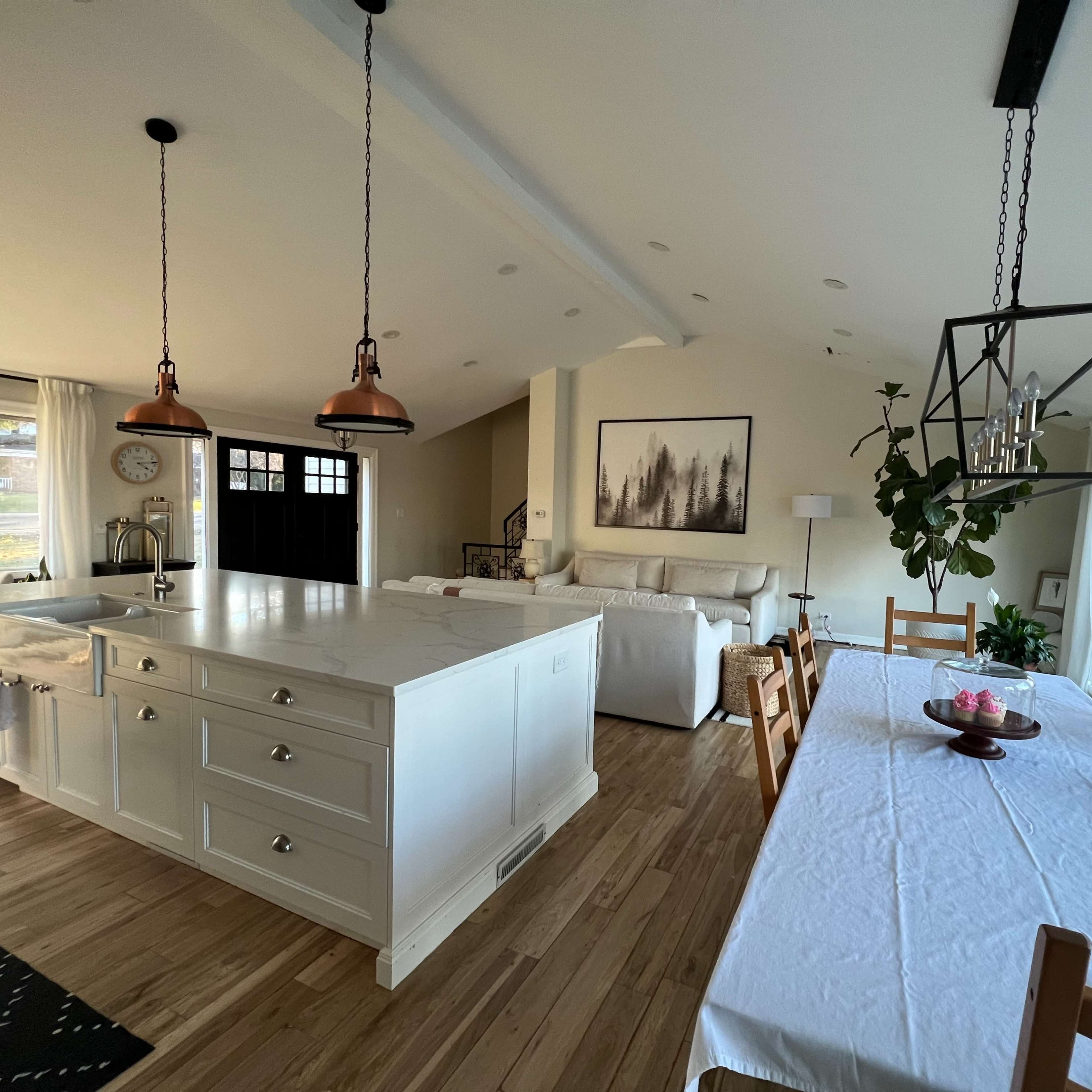 An open-concept kitchen and dining area featuring a large island with a white countertop, wooden dining table, and modern lighting fixtures.